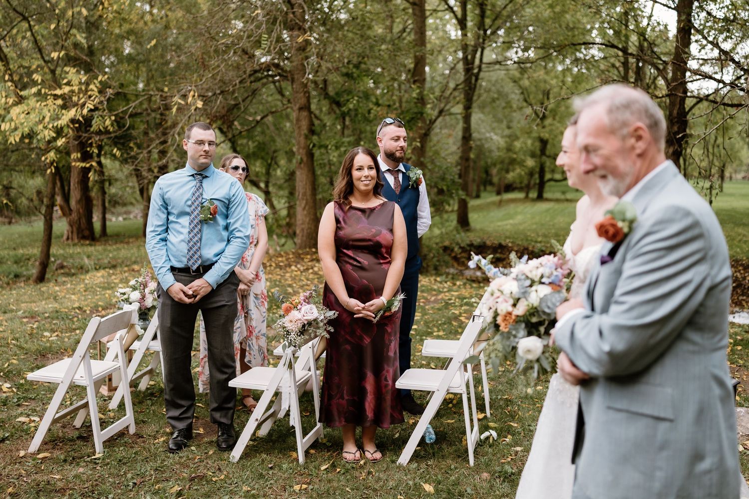 Wedding guests watch emotional outdoor ceremony with white folding chairs and floral arrangements in wooded setting.