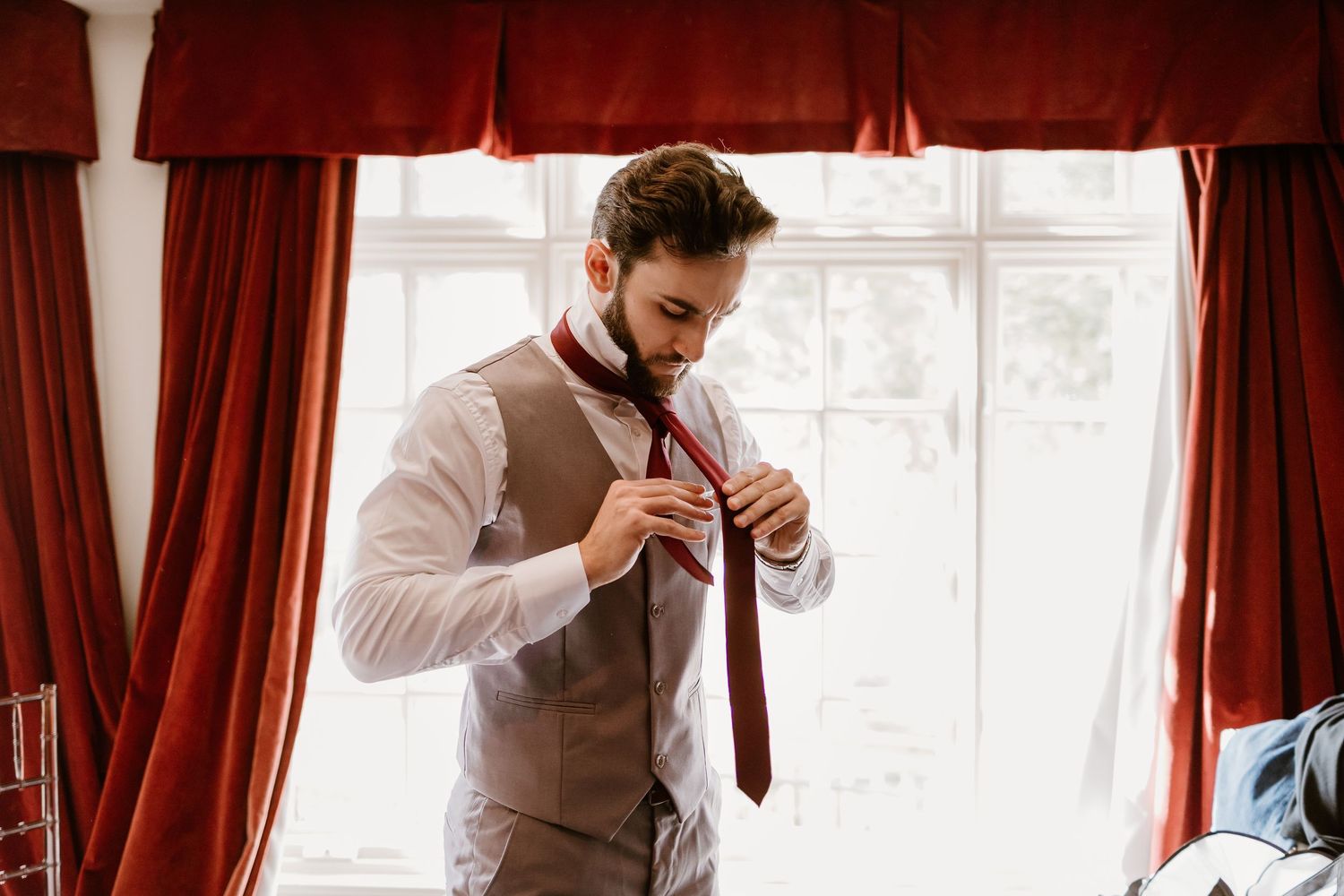 Person in beige vest adjusts red tie while getting ready by window.