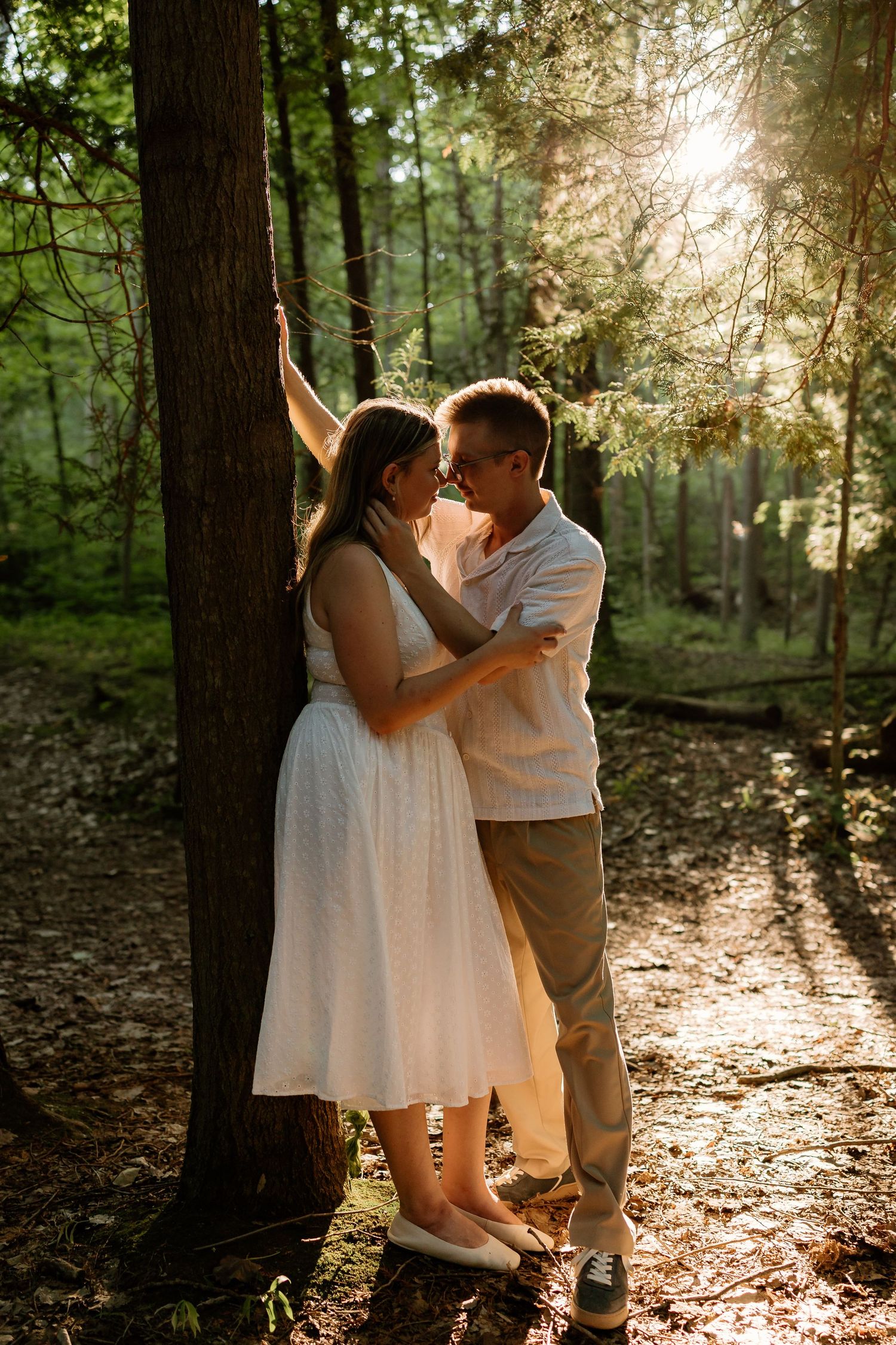 Couple sharing a romantic moment by a tree in a sunlit forest, woman in white dress and man in casual attire.