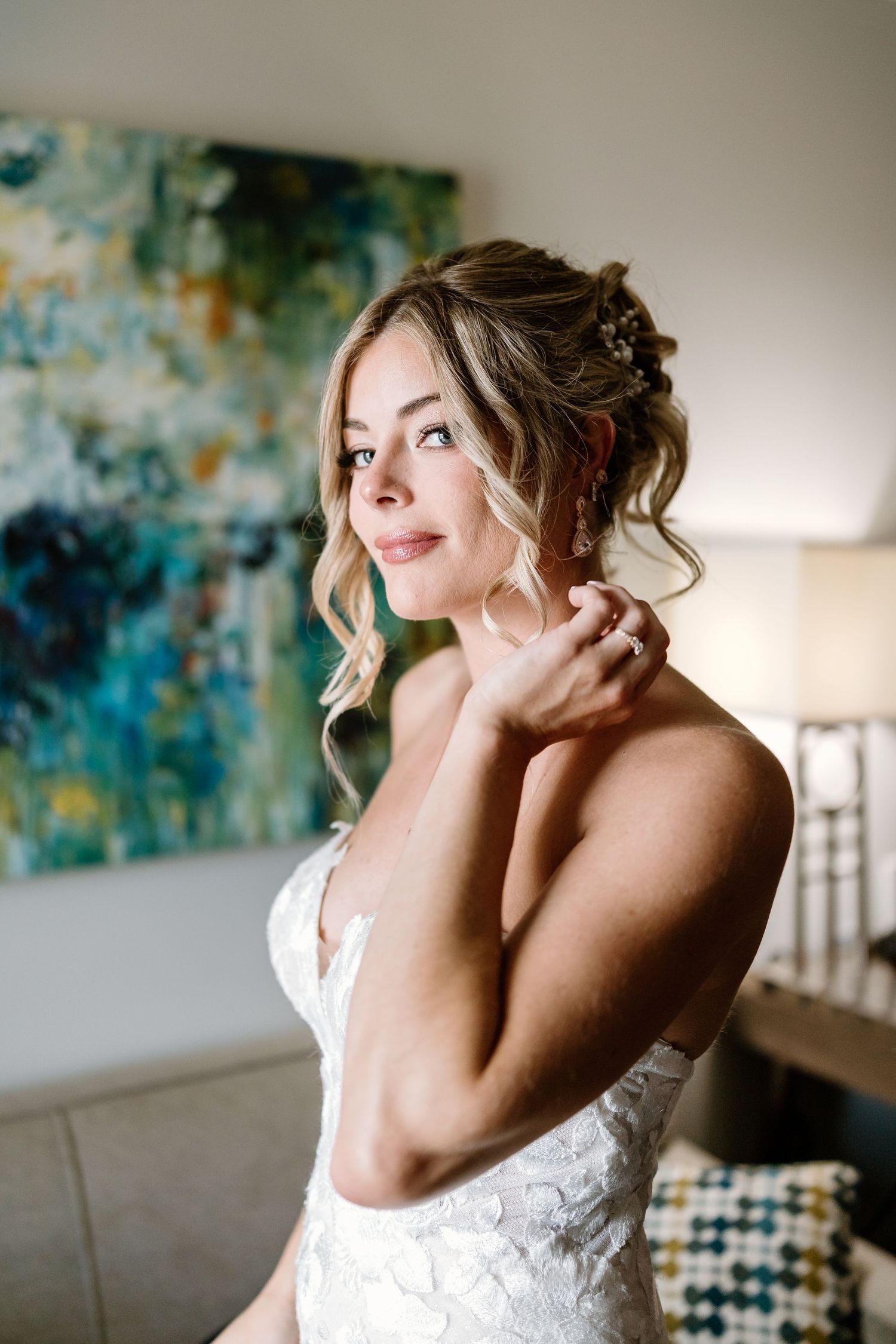 Bride in lace wedding dress adjusting earring, elegant updo with floral pins, soft natural makeup, colorful abstract art background.