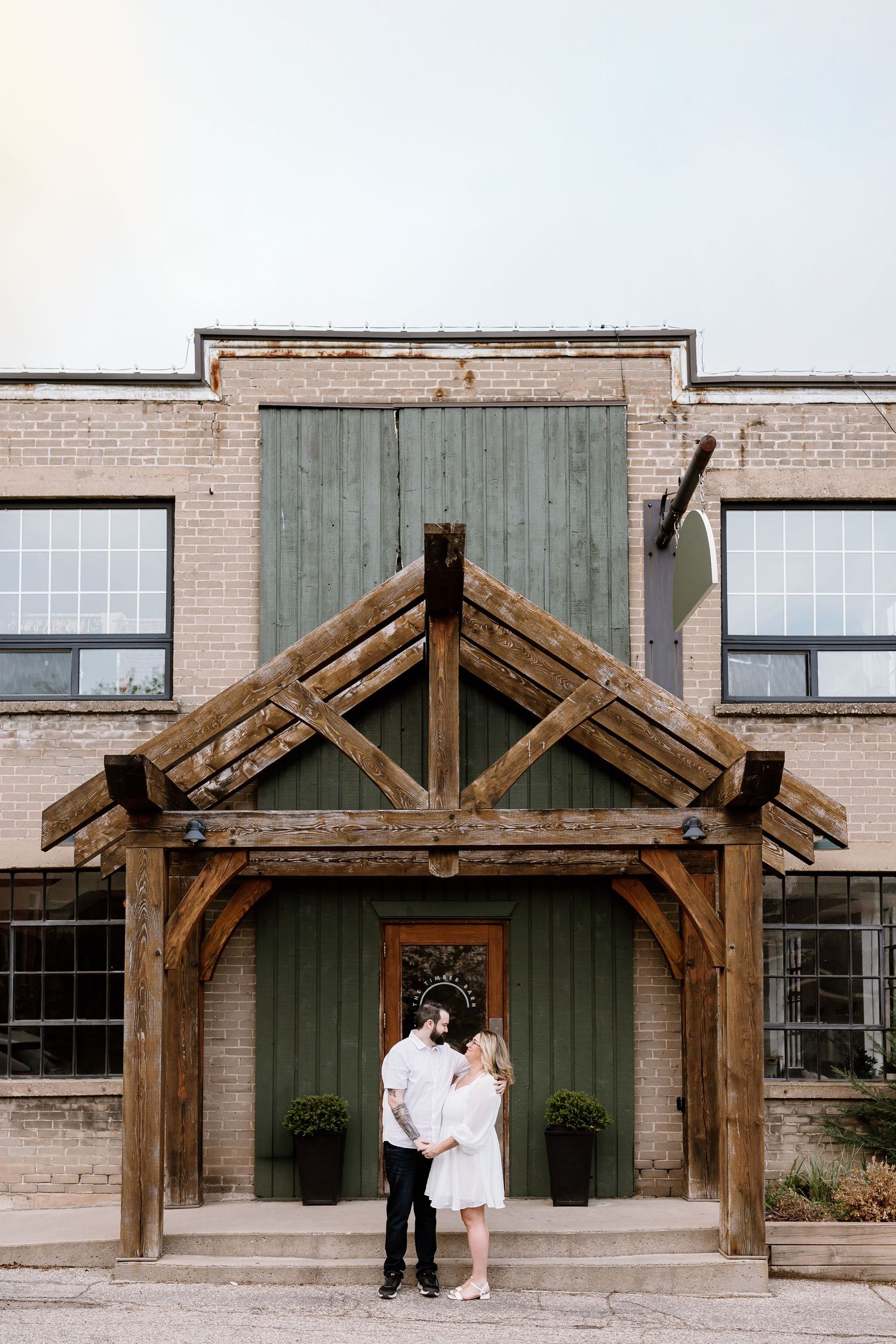 Couple embraces under a wooden timber frame entrance of a rustic brick wedding venue.
