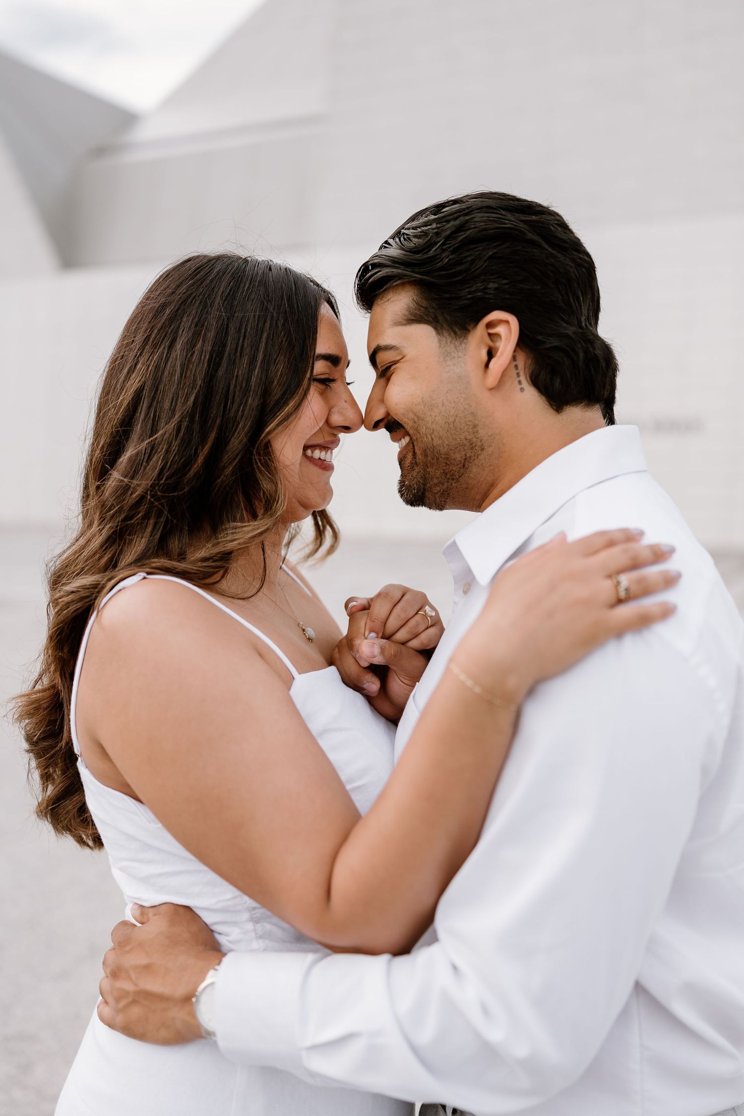 Happy couple in white outfits sharing a romantic moment, smiling and holding hands outdoors.