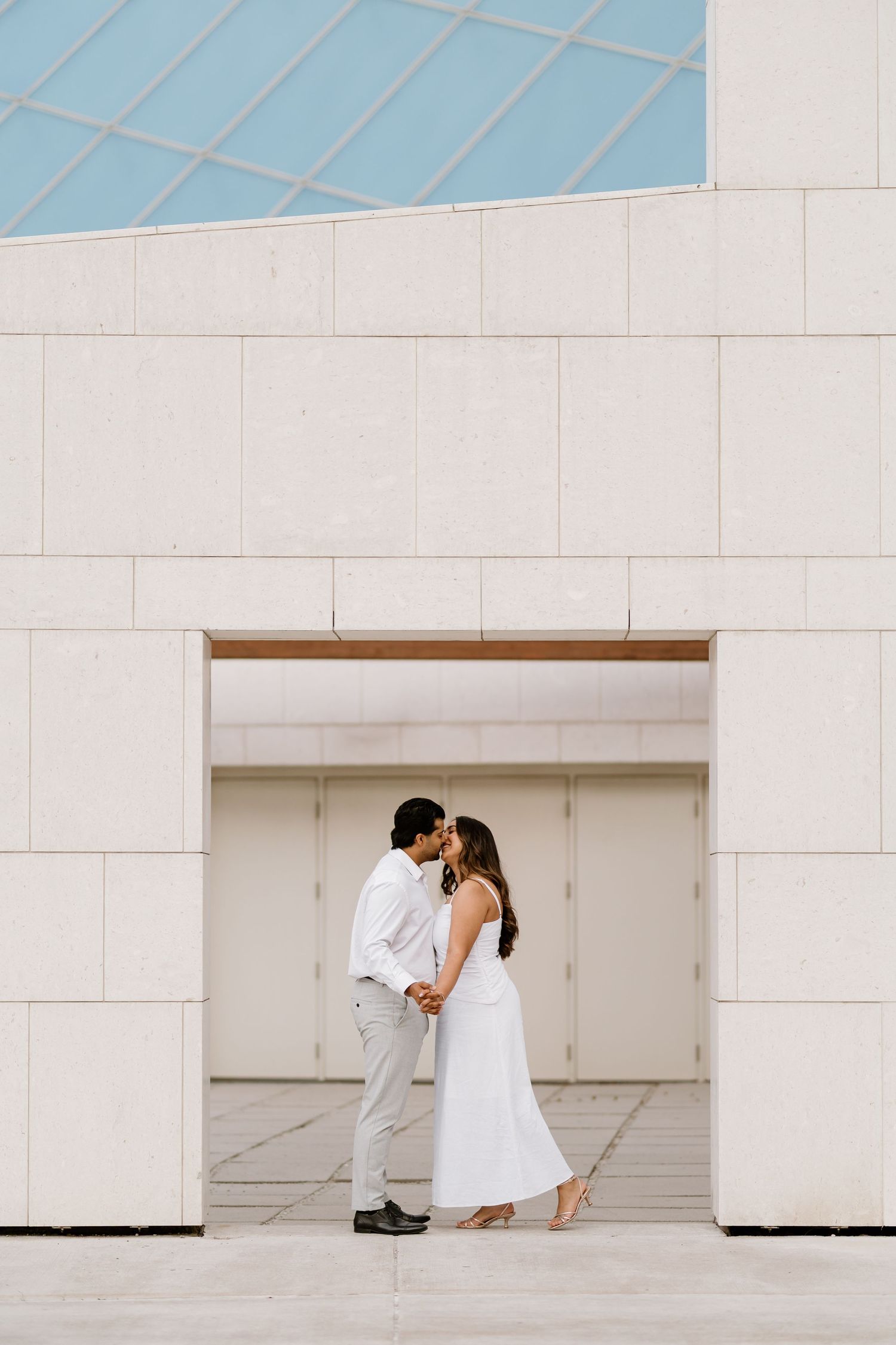 Couple sharing a romantic kiss in white outfits framed by a modern white stone building entrance.