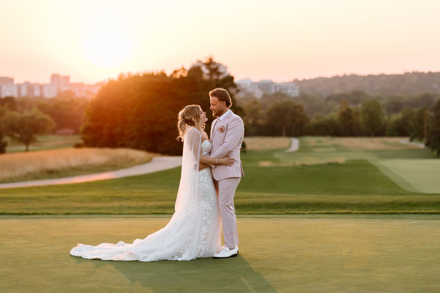 Bride and groom embrace on a golf course at golden hour sunset with lush green fairways and city skyline in background.