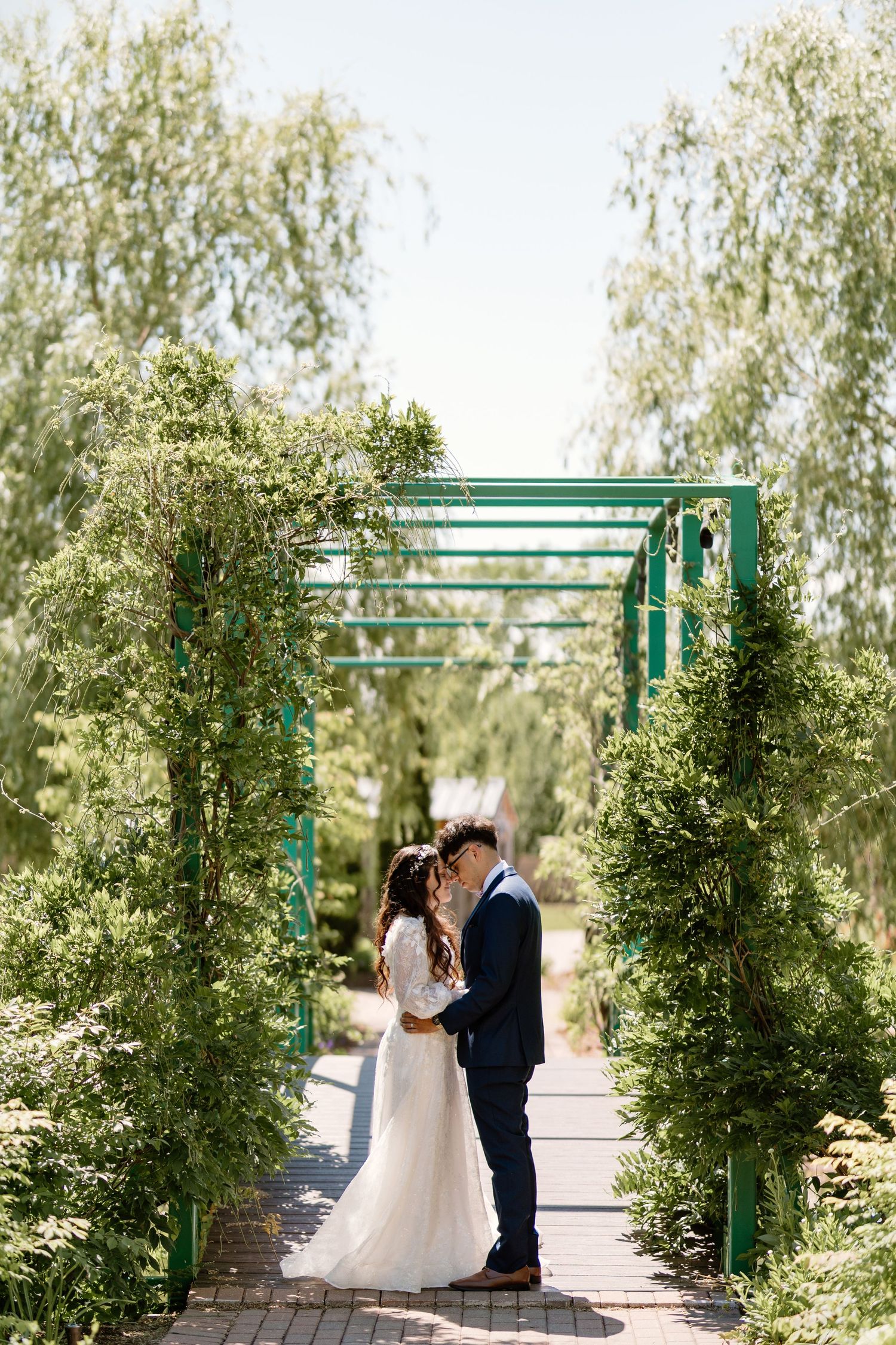 Bride and groom share a romantic moment under a green garden arch surrounded by lush trees on their wedding day.