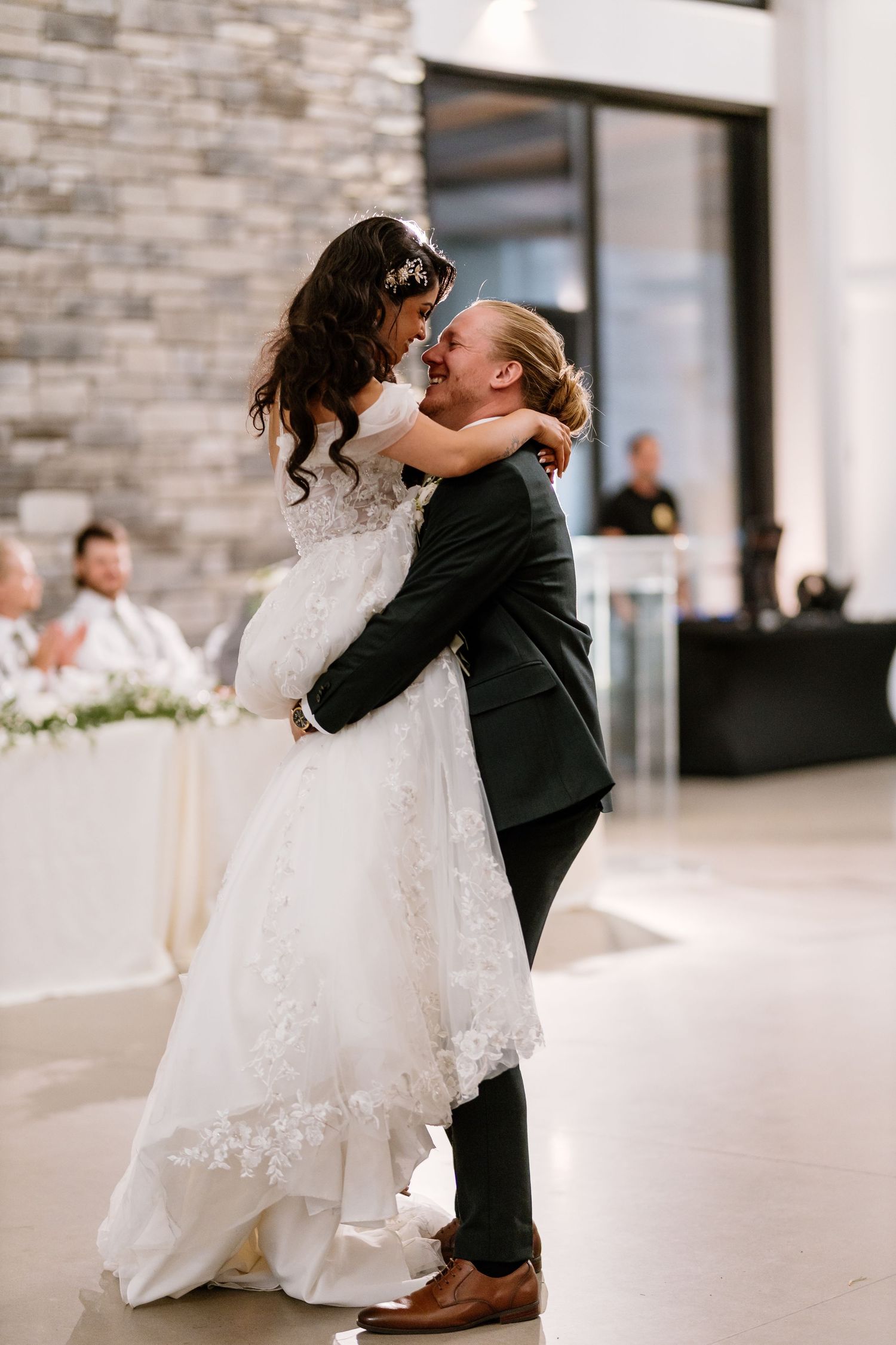 Groom lifts bride in white lace wedding dress during romantic first dance at elegant indoor wedding reception.