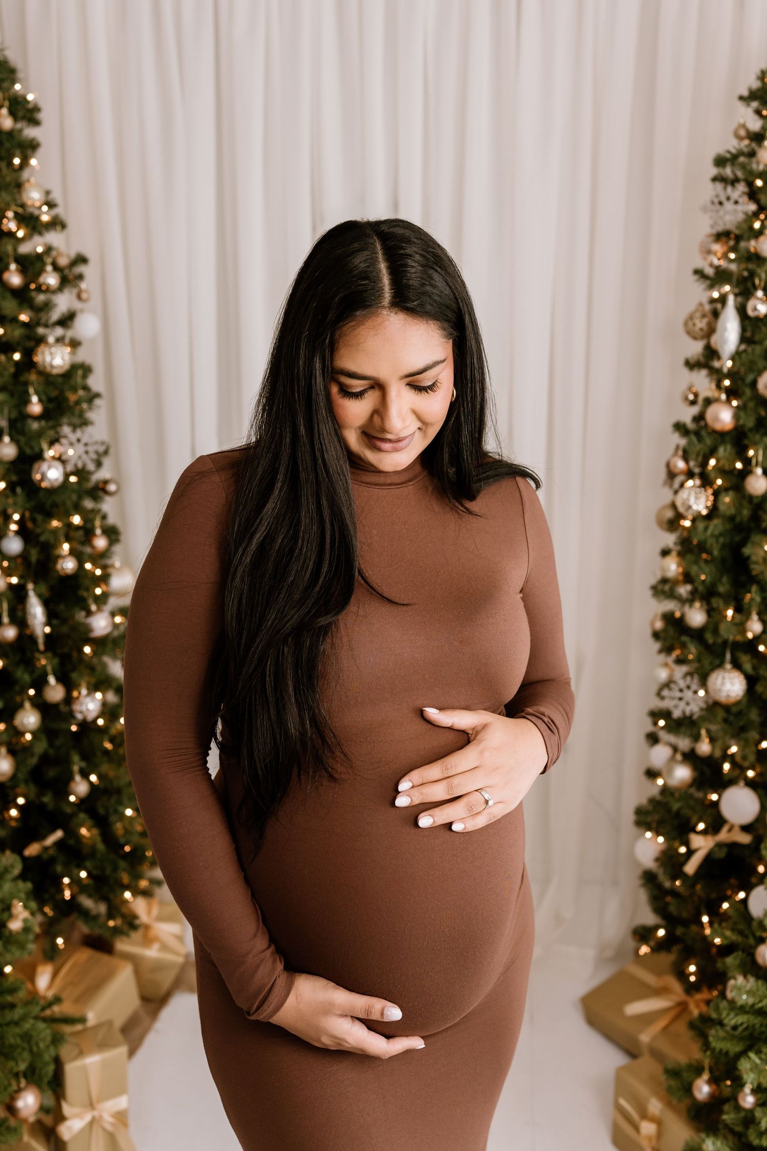 Maternity photos against white curtain and Christmas trees with gold decorations.