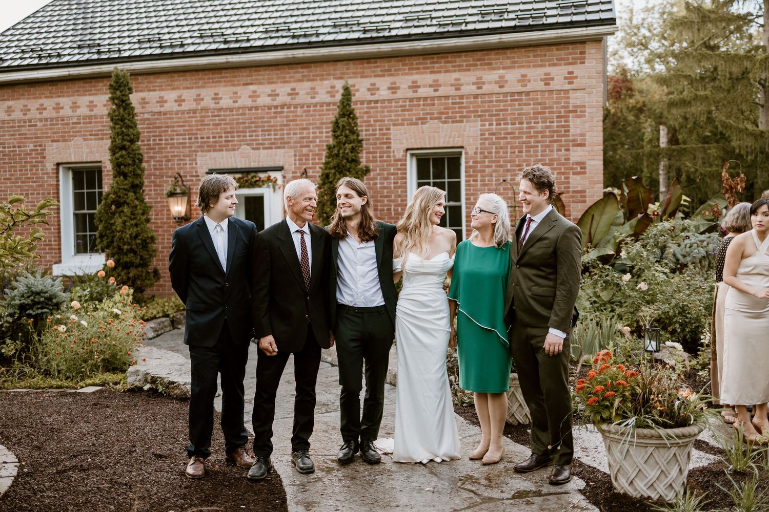 A wedding party poses for a photo outside a brick building with landscaped gardens.