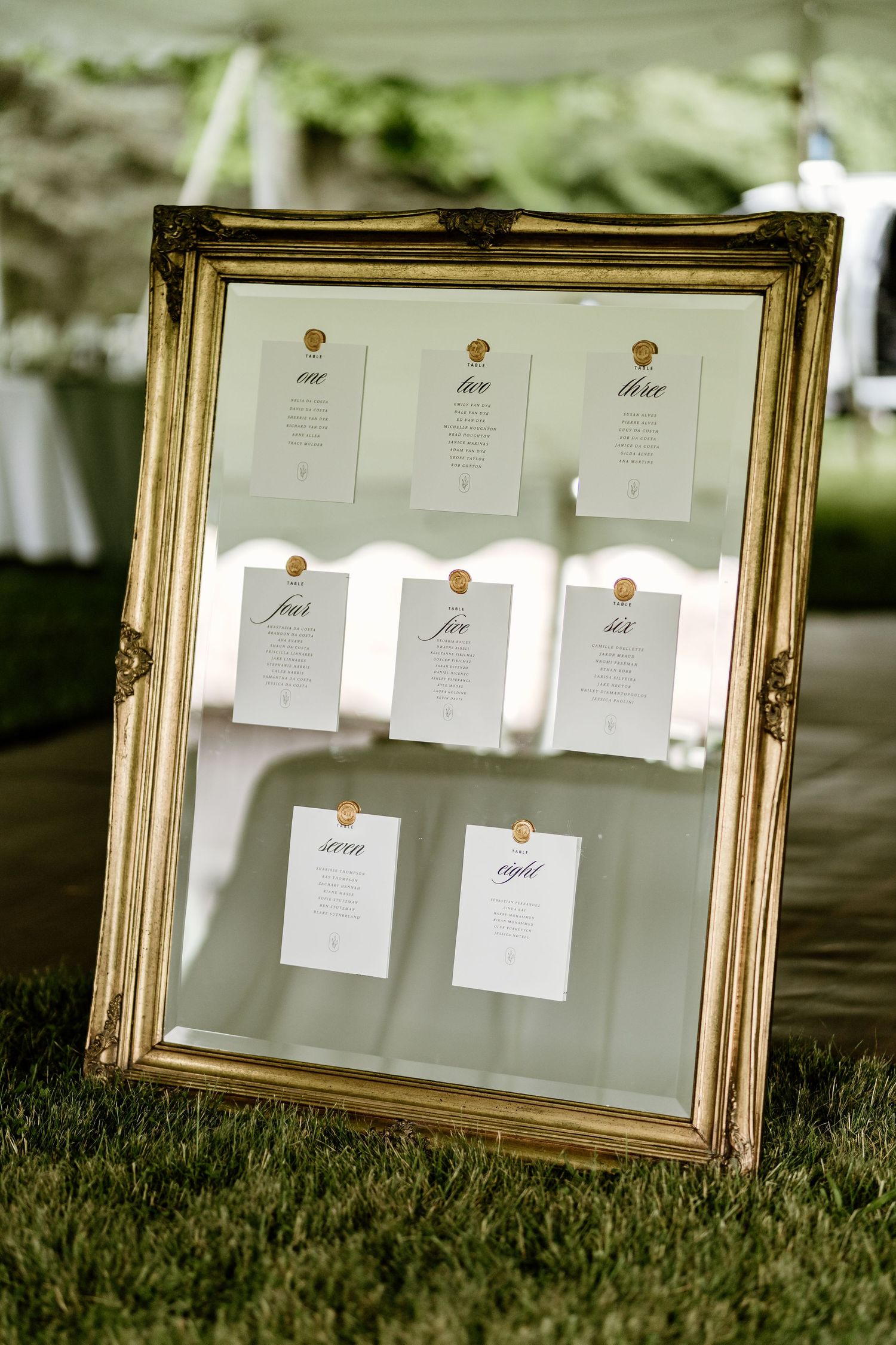Gold framed seating chart display with white cards arranged on grass at an outdoor wedding event.
