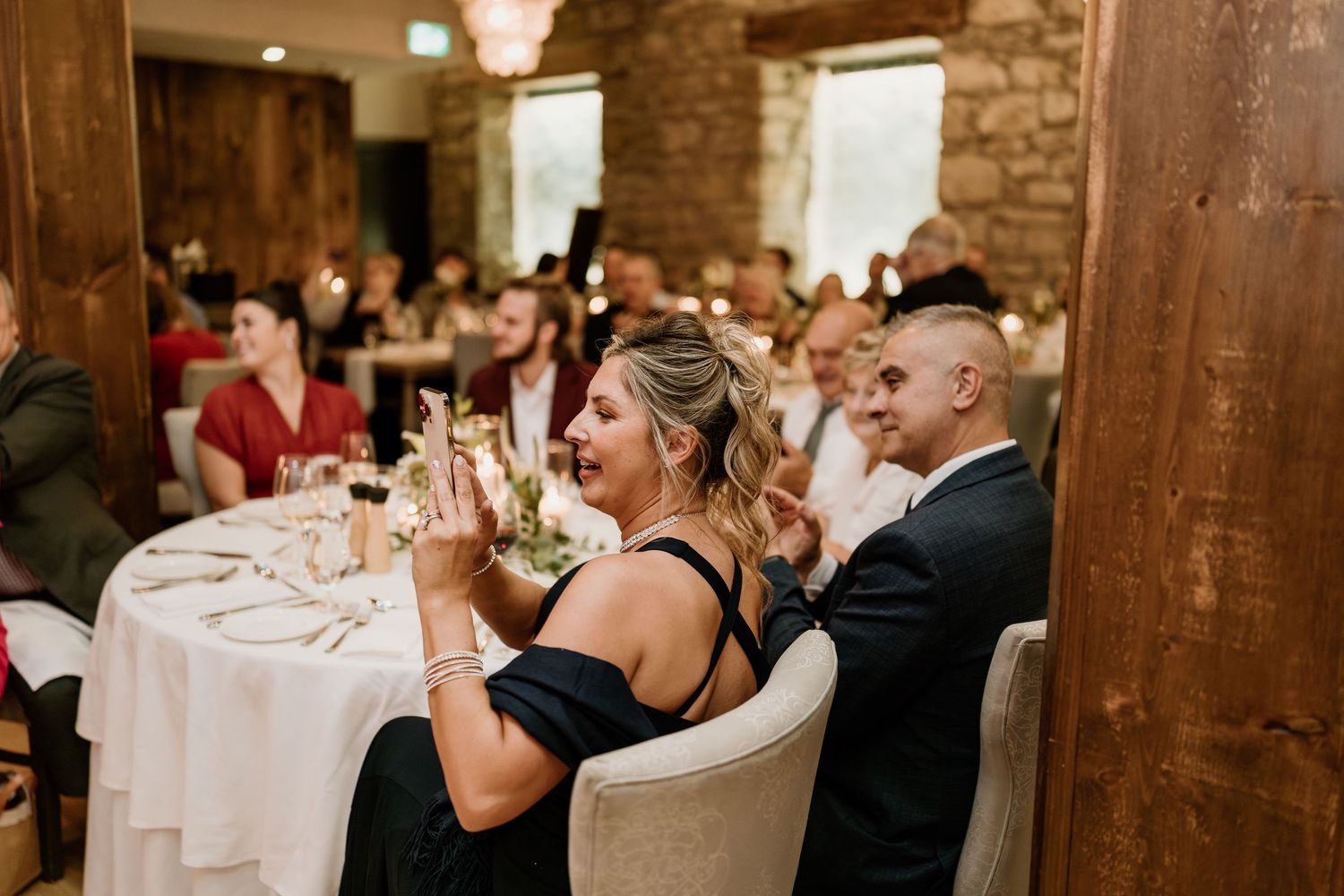 Wedding reception guests seated at elegant white-clothed tables in a stone-walled venue with chandeliers.