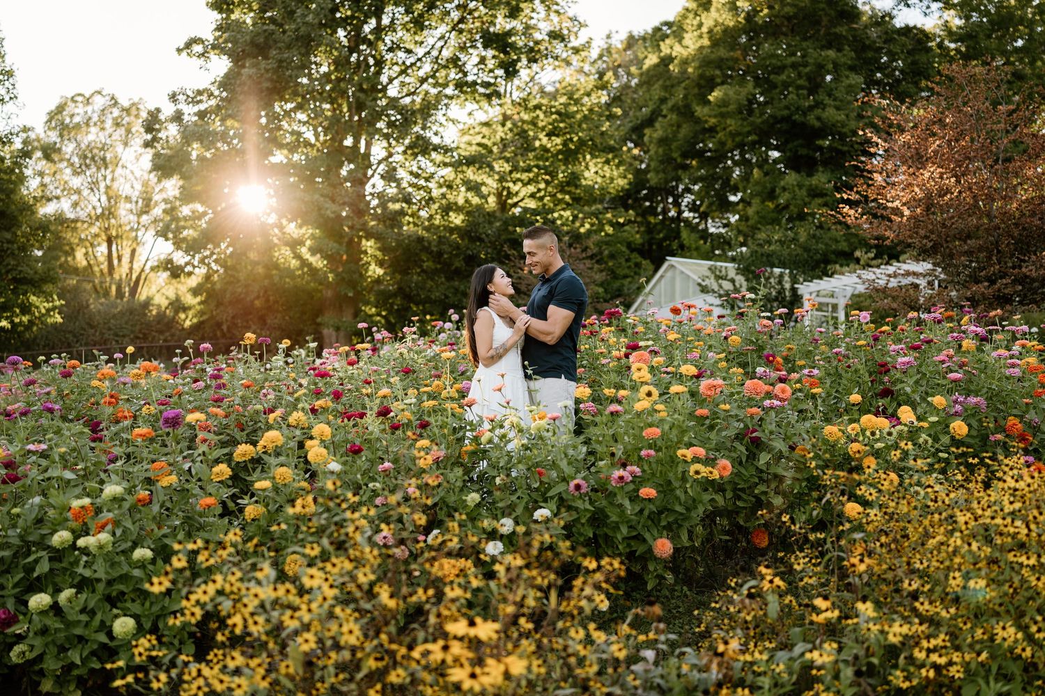 Couple embracing in a colorful wildflower garden at sunset, with warm golden sunlight filtering through tall trees in the background.