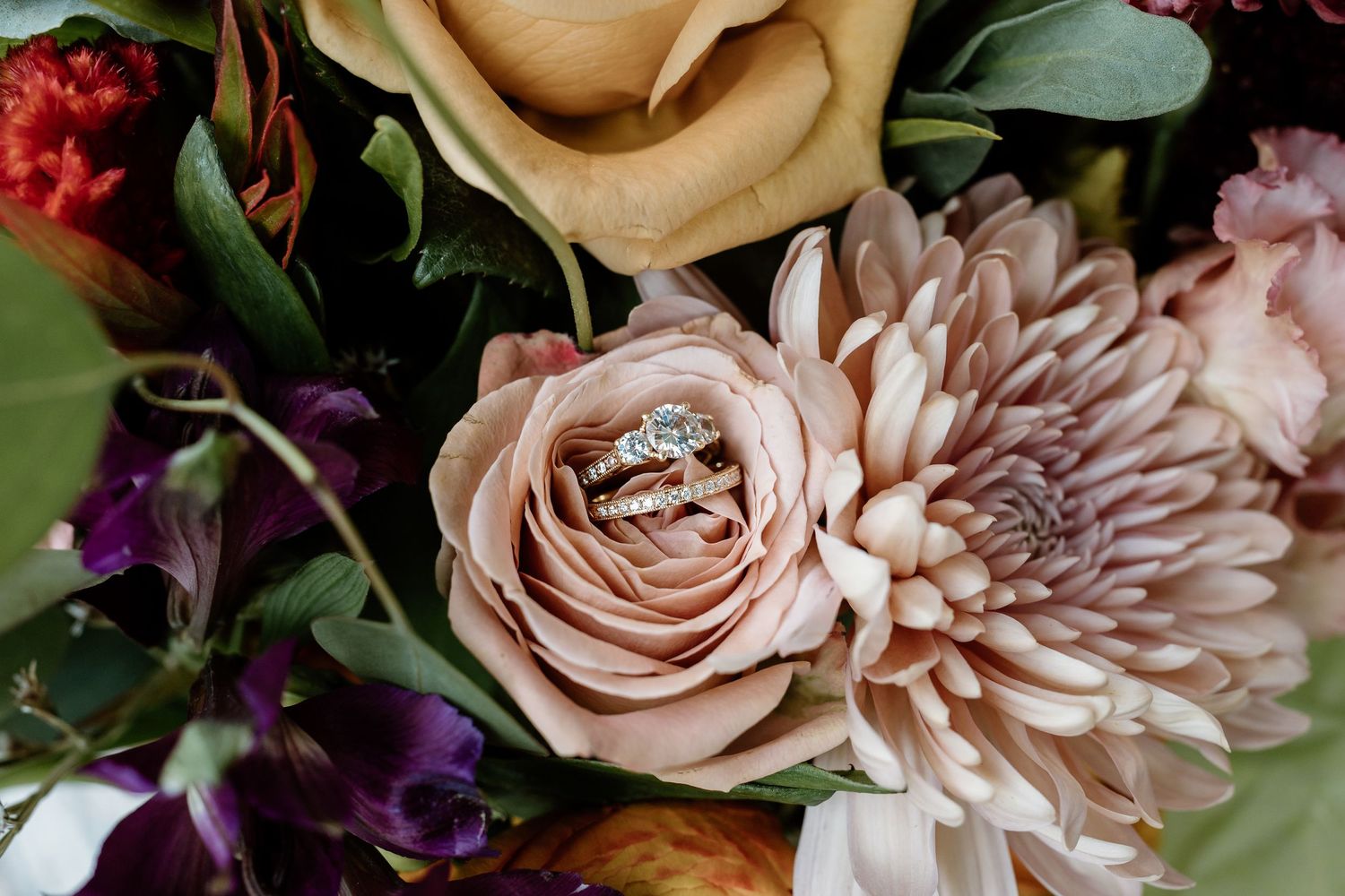 Close-up of wedding rings nestled in a blush pink rose among a lush floral arrangement.