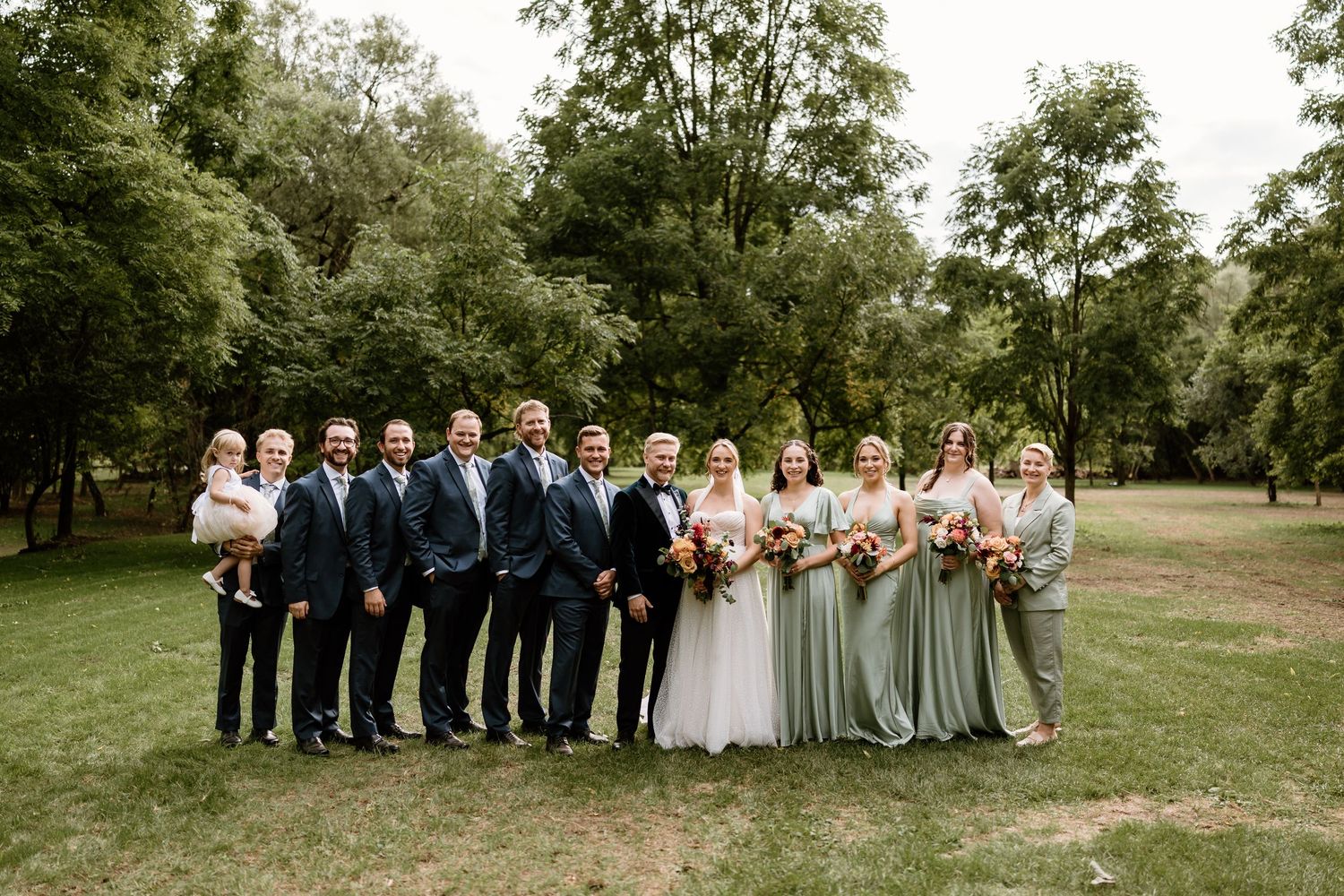Wedding party members lined up in formal attire with bridesmaids in sage green dresses.