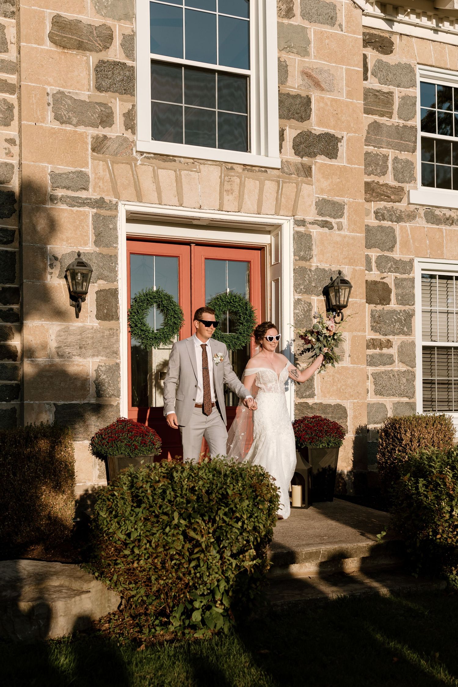 Wedding party poses in front of a rustic stone building with pink doors and greenery.