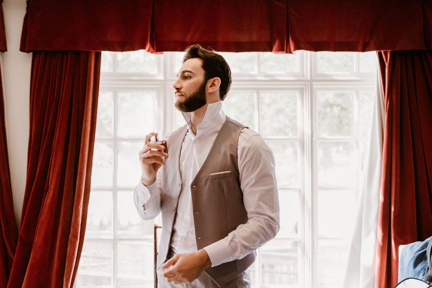 Person adjusts tie while getting ready by window with red curtains.