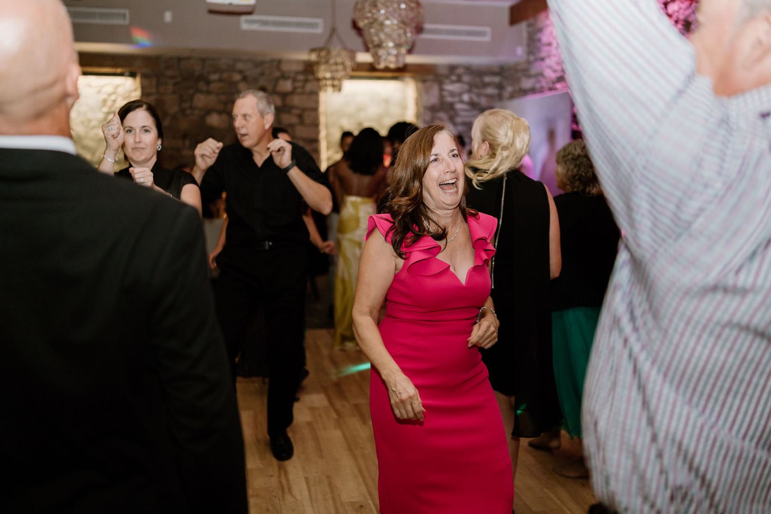 Wedding guests enjoying themselves on dance floor with pink lighting and stone walls.