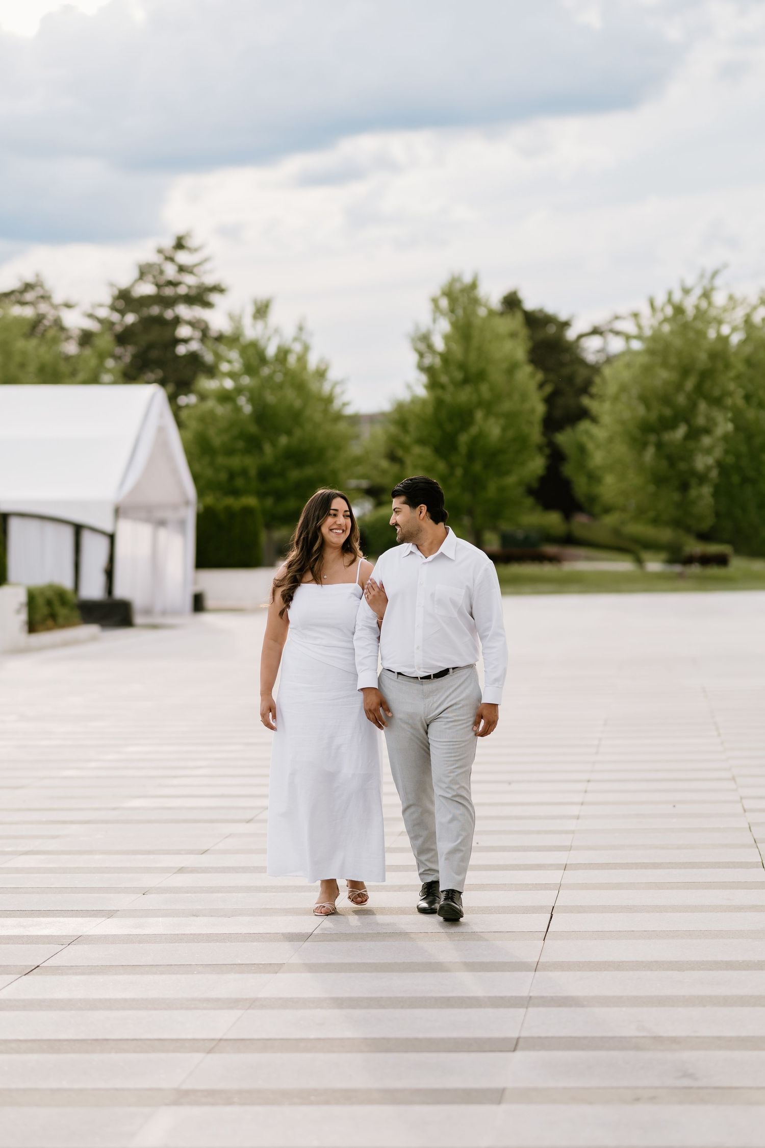 Couple in white outfits walking hand in hand on a paved path surrounded by lush green trees.