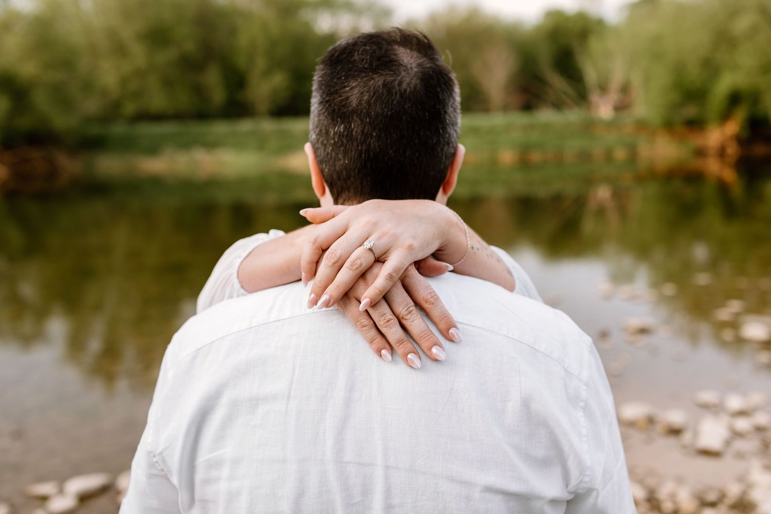 Couple embracing by a lake, woman's hand with engagement ring resting on man's shoulder in nature.