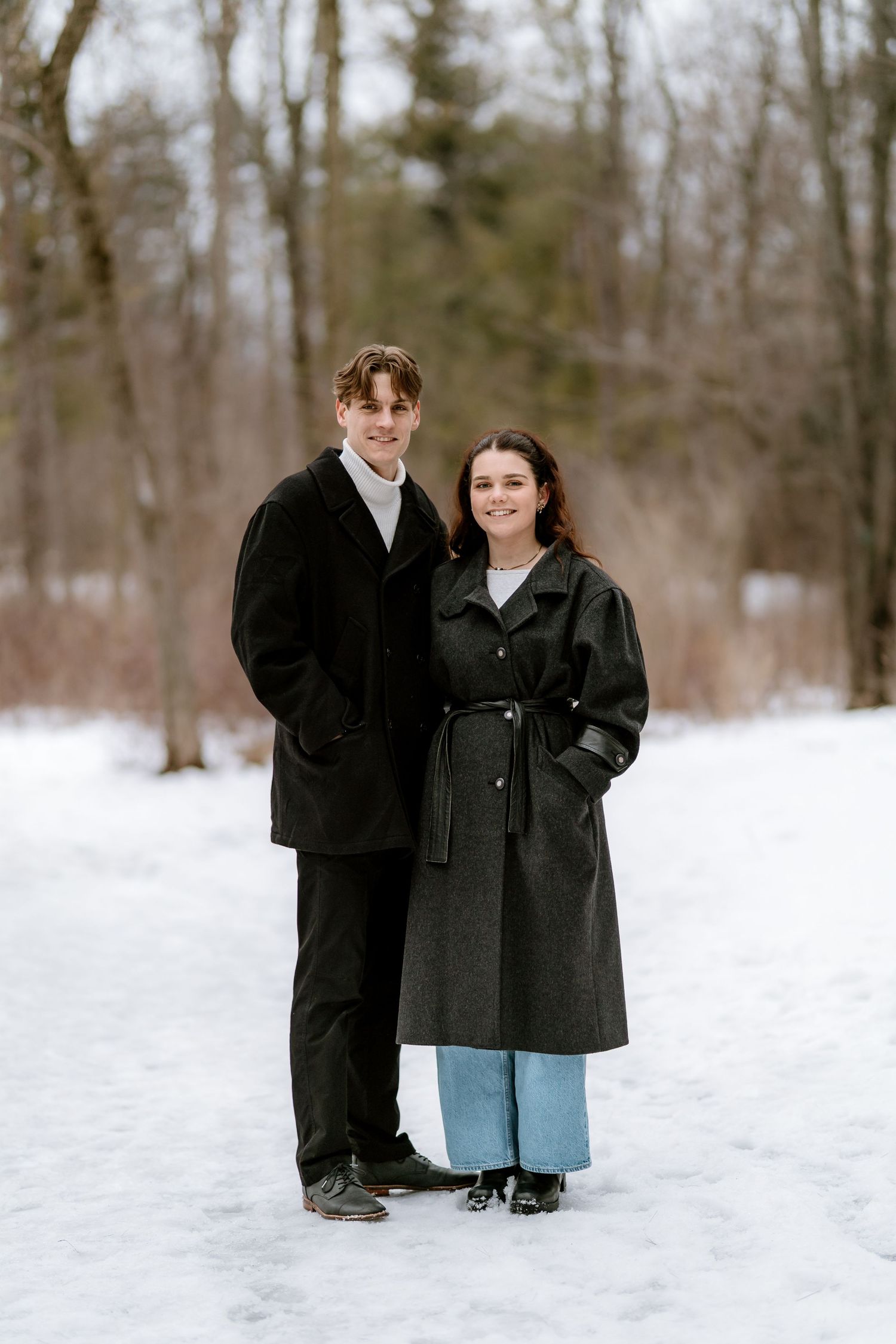 A couple in matching black winter coats stands together in the snow.