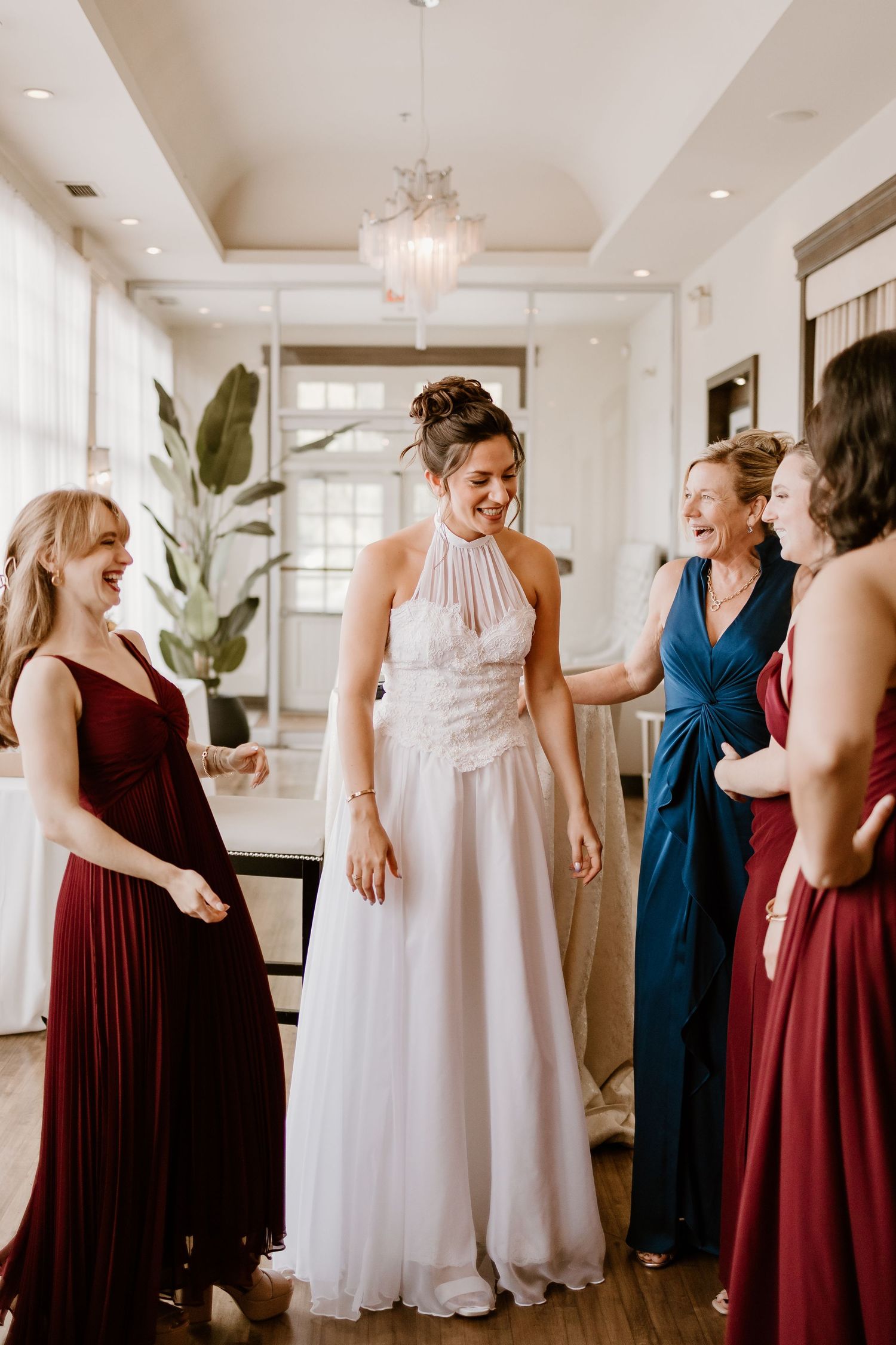 Wedding party shares joyful moment in a bright room with elegant chandelier and potted plants.