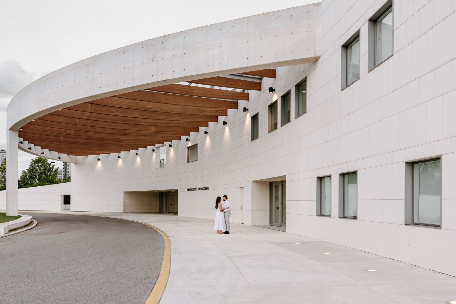 Couple embracing outside a modern curved white building with wooden canopy and elegant architecture.