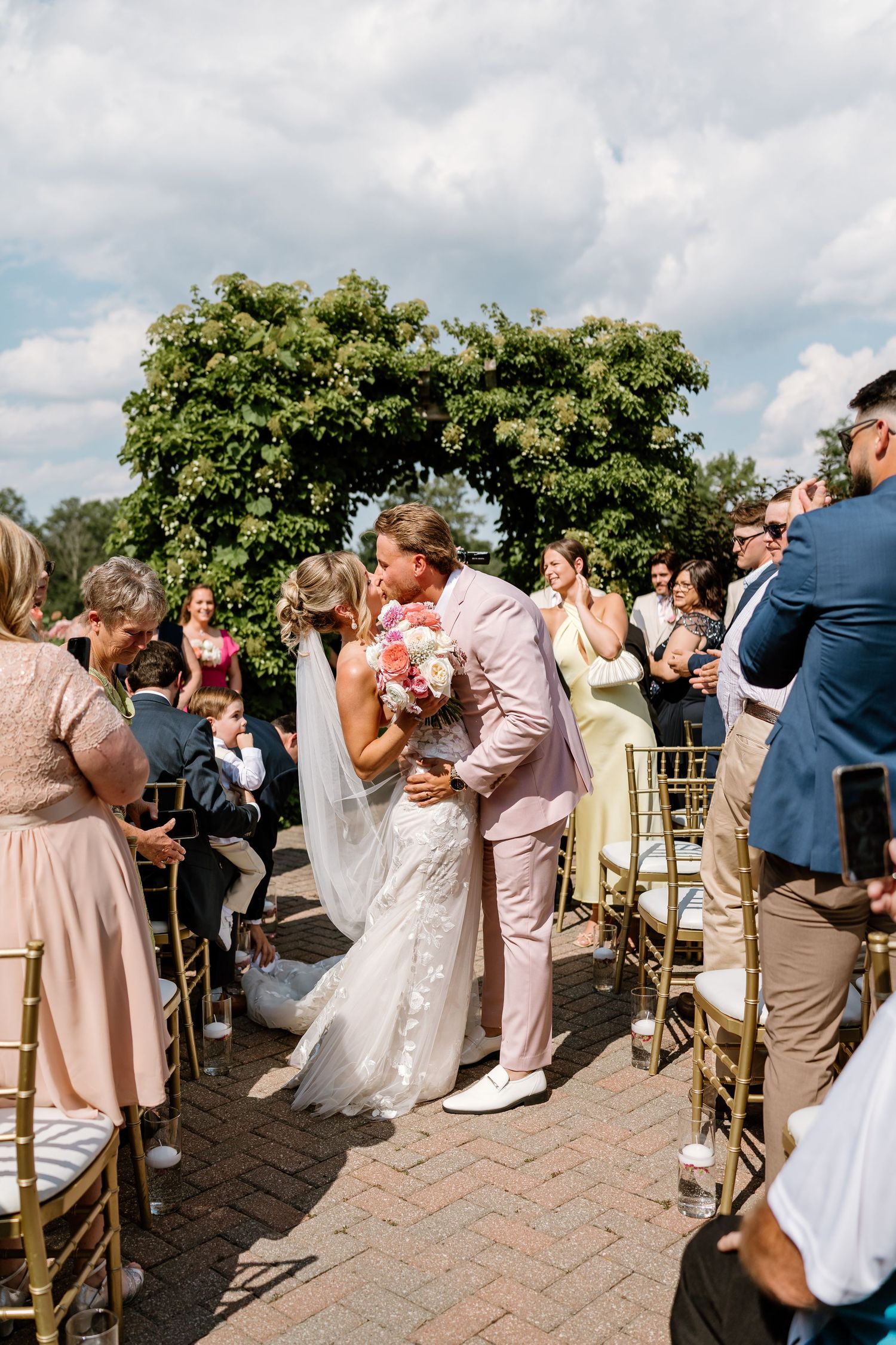 Bride and groom share first kiss as newlyweds during outdoor wedding ceremony with guests celebrating.