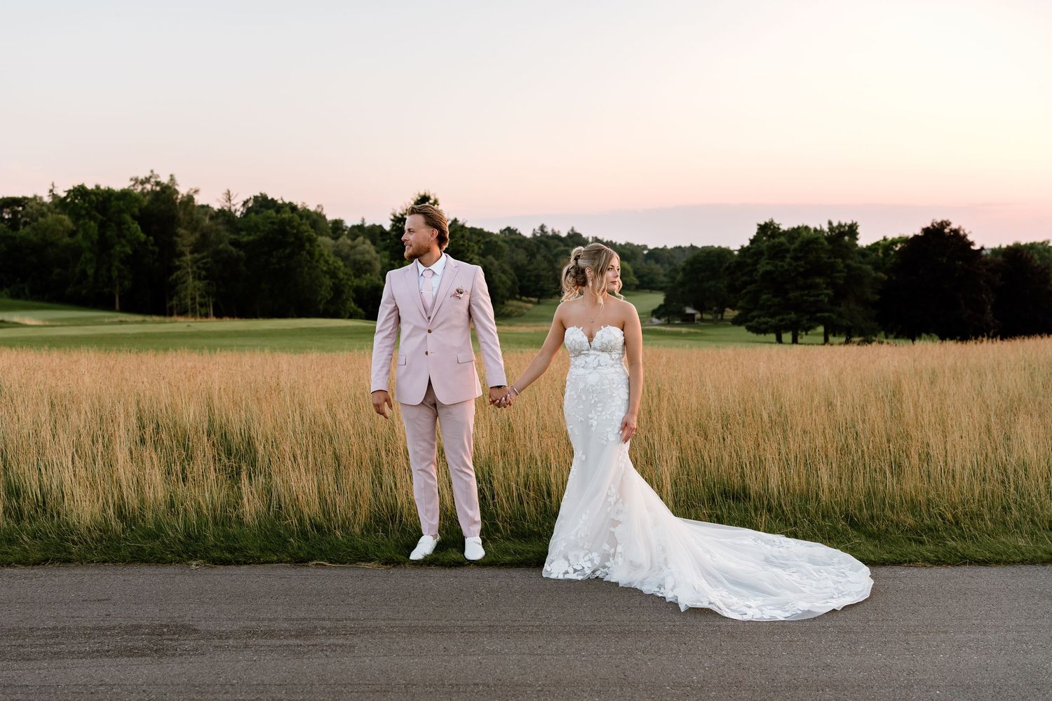 Bride in lace gown and groom in pink suit hold hands at sunset on a golf course path.