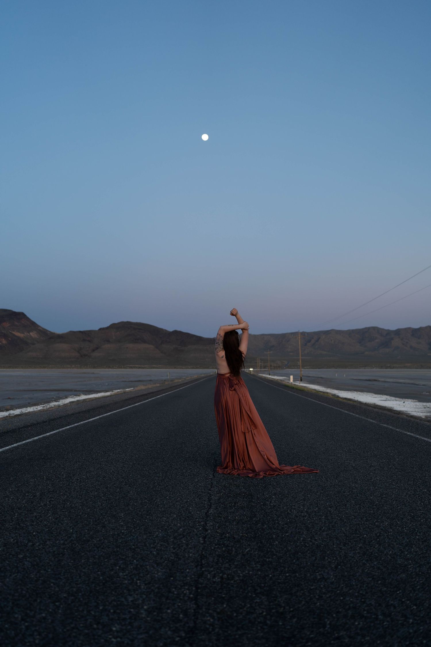 A person in a flowing burgundy dress stands on an empty desert highway under a full moon and twilight sky.