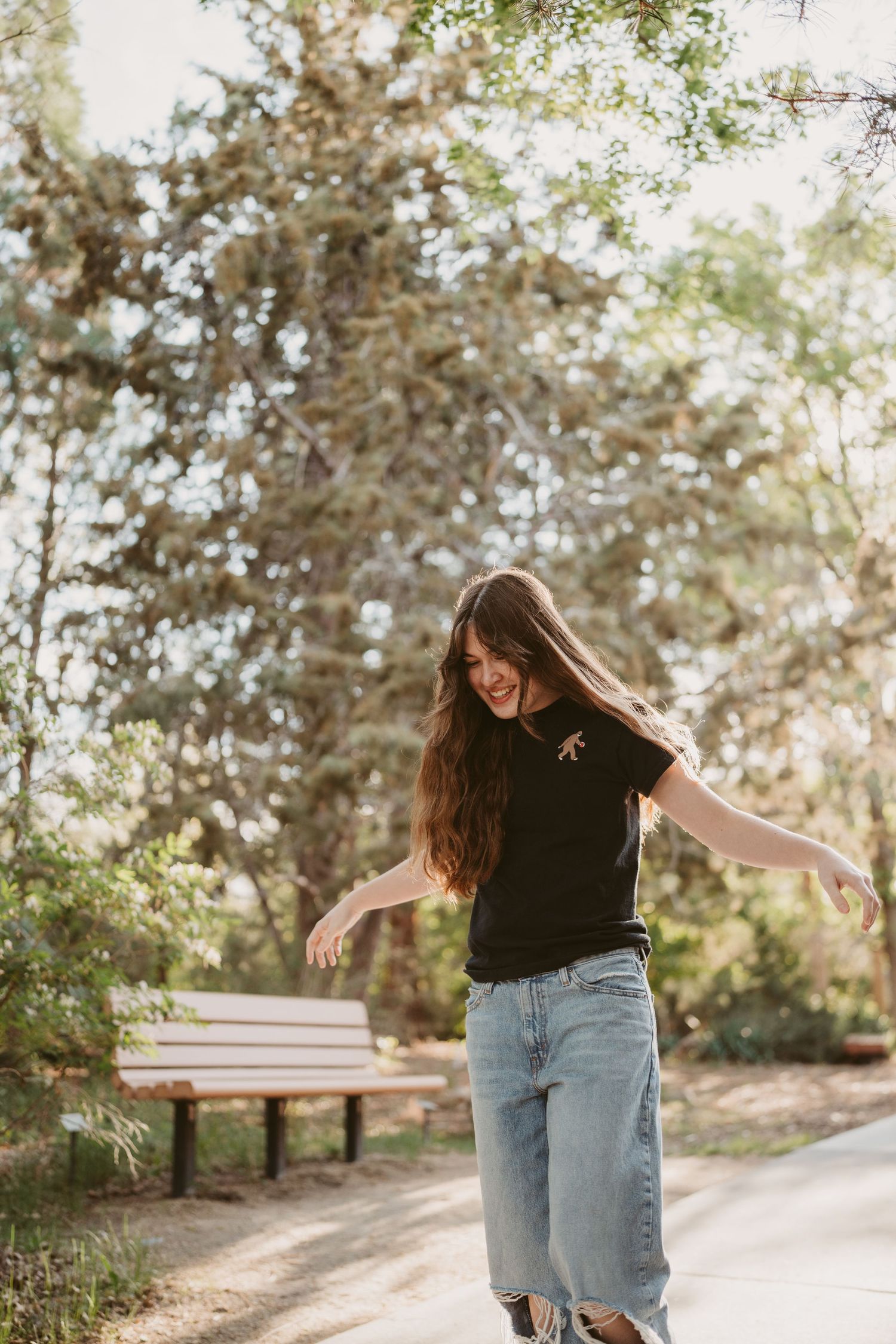 A person in a black shirt and jeans dances joyfully on a sunlit path surrounded by trees.