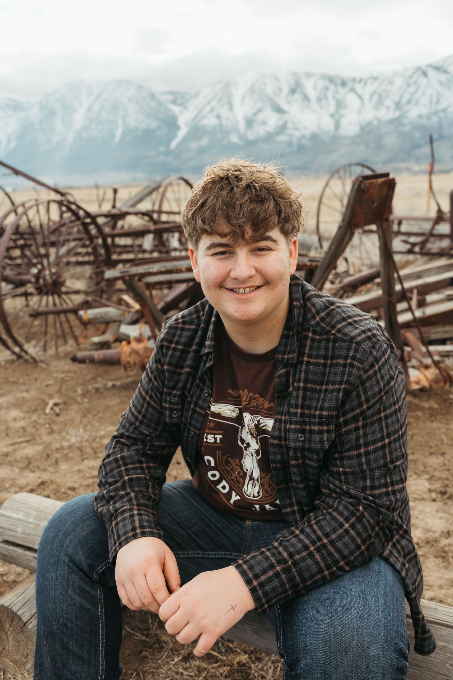 A person in a plaid shirt and jeans sits casually outdoors with snow-capped mountains visible in the background.