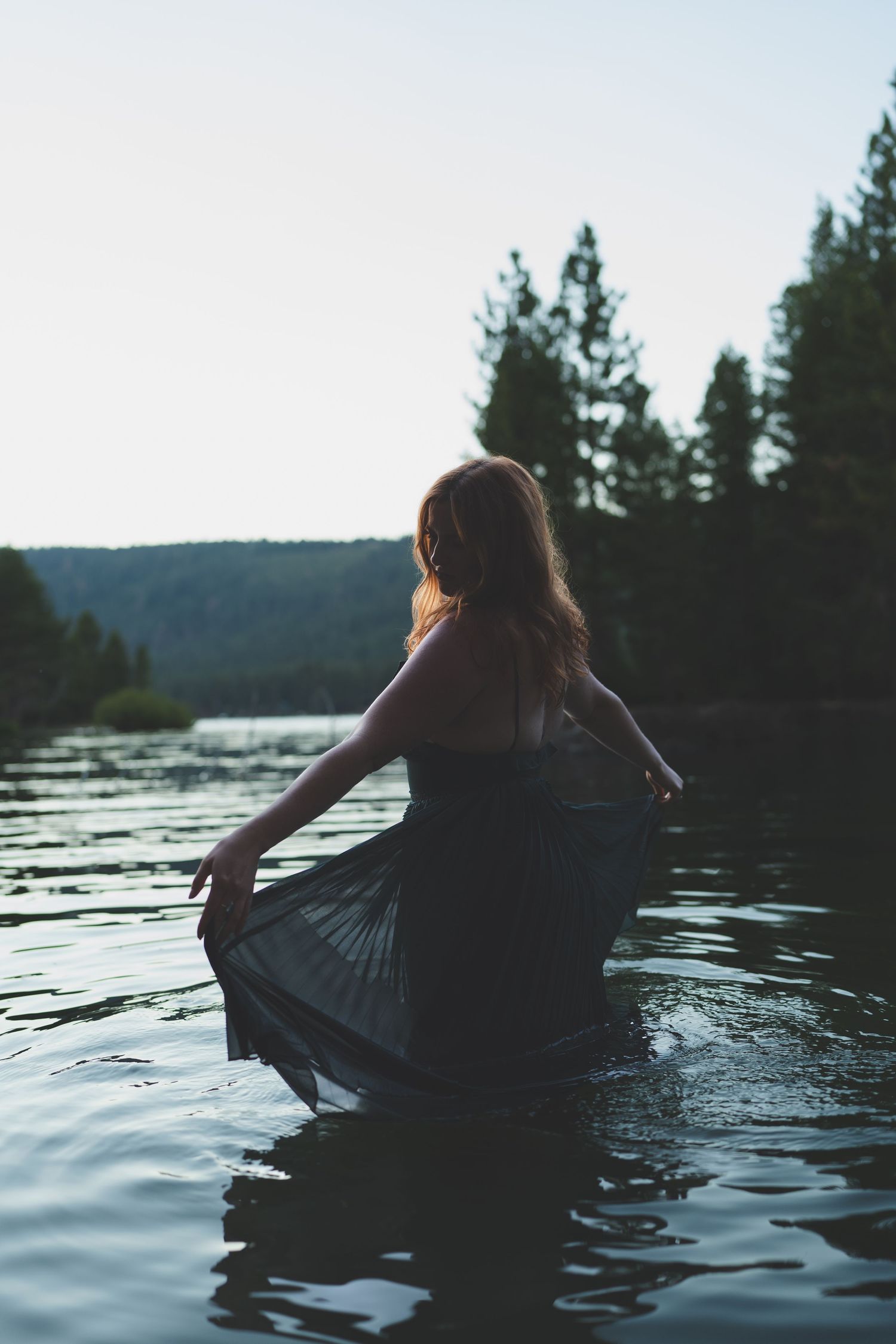 A woman in a dark flowing dress stands waist-deep in a serene lake surrounded by pine trees at dusk.