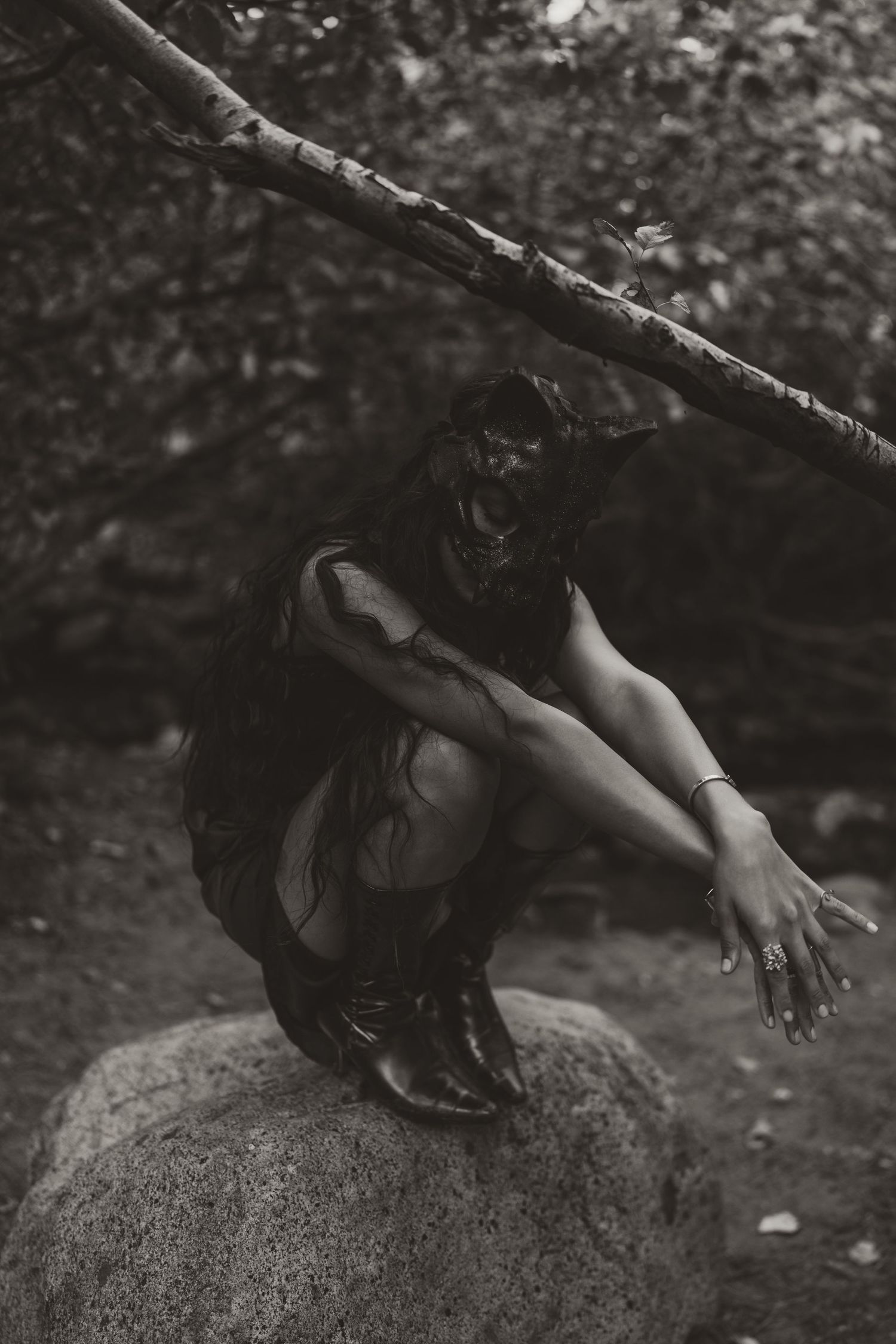 Black and white photo of a woman in dark dress and mask crouching on a rock in a moody forest setting.