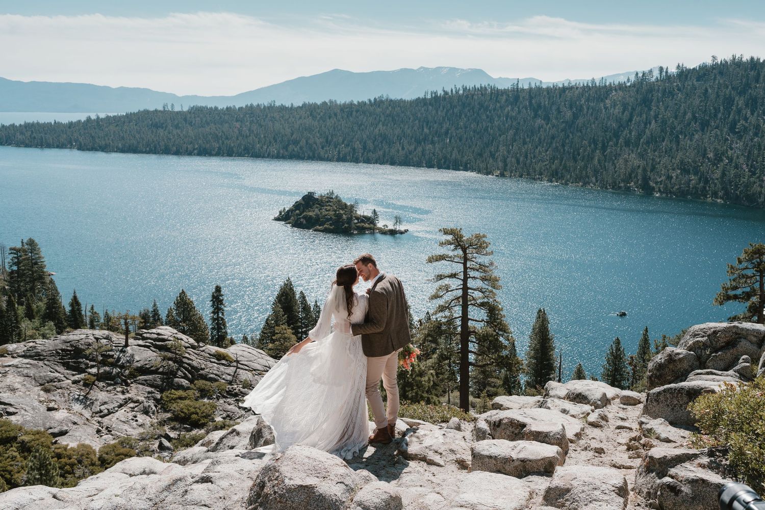 Couple shares a kiss at Emerald Bay, Lake Tahoe, with stunning blue water, forested island, and mountain views behind them.