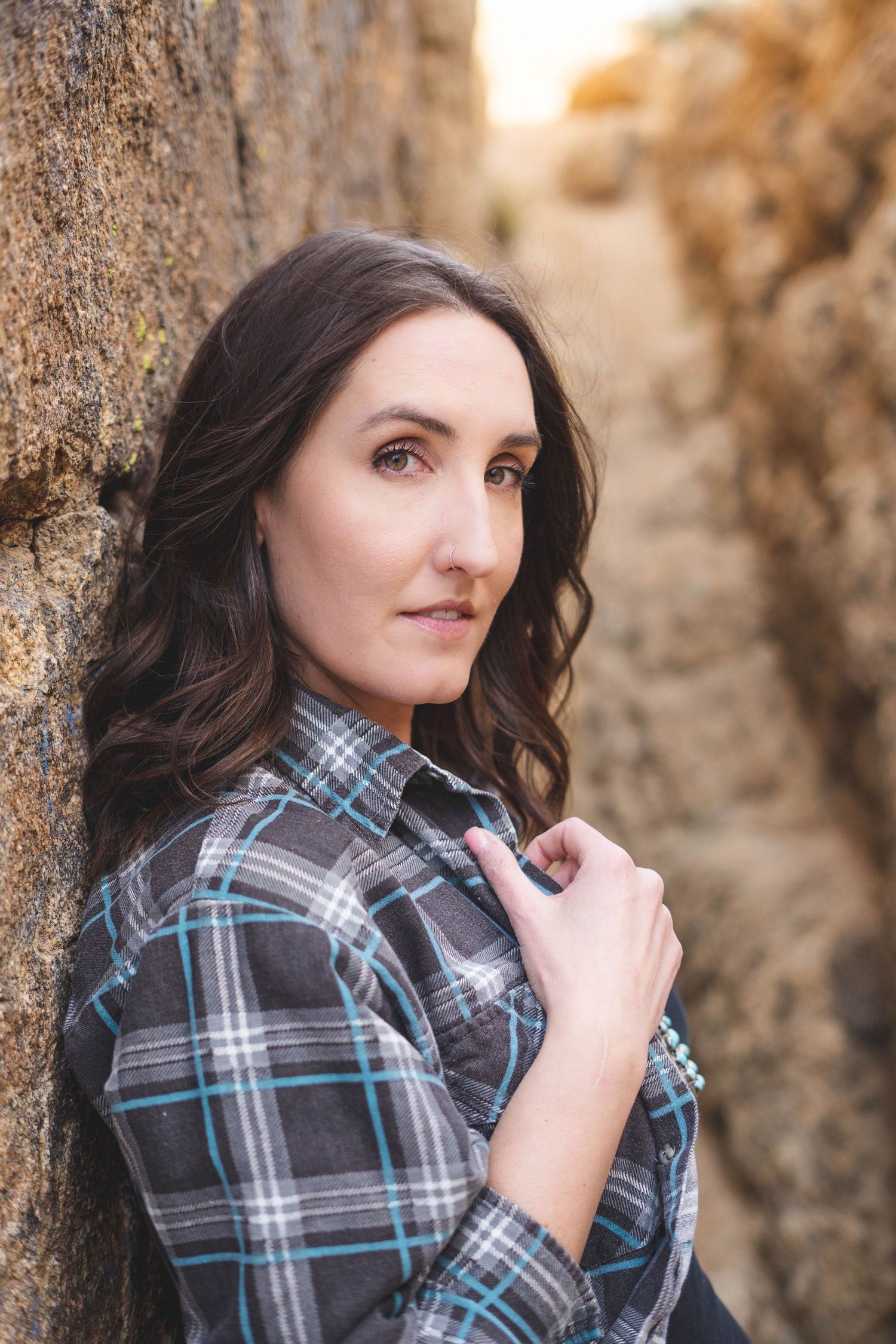 A young woman with long dark wavy hair leans against a rocky cliff wearing a gray and teal plaid flannel shirt.