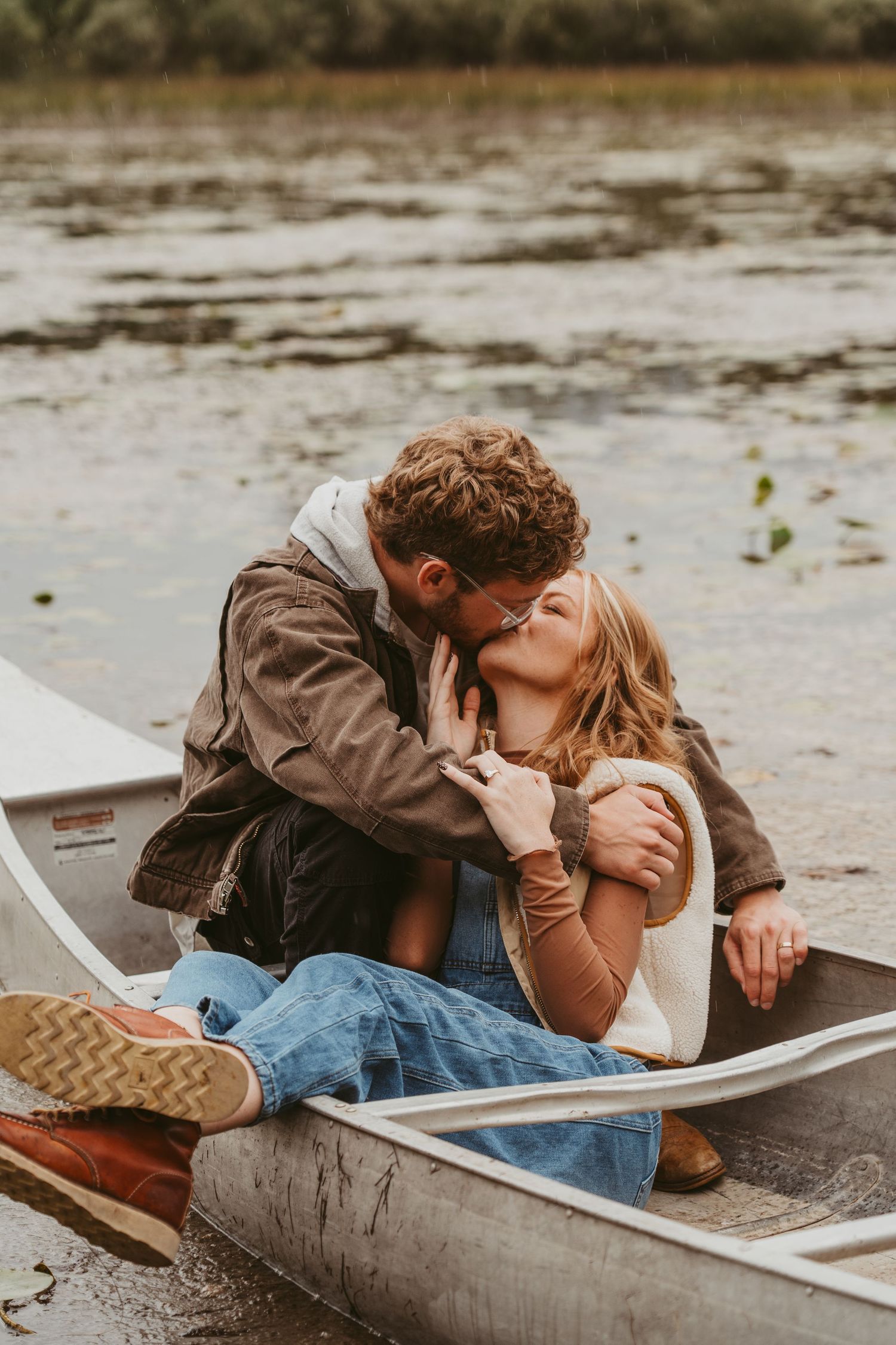 A romantic couple sharing intimate moments while sitting in a canoe by the water on an overcast day.