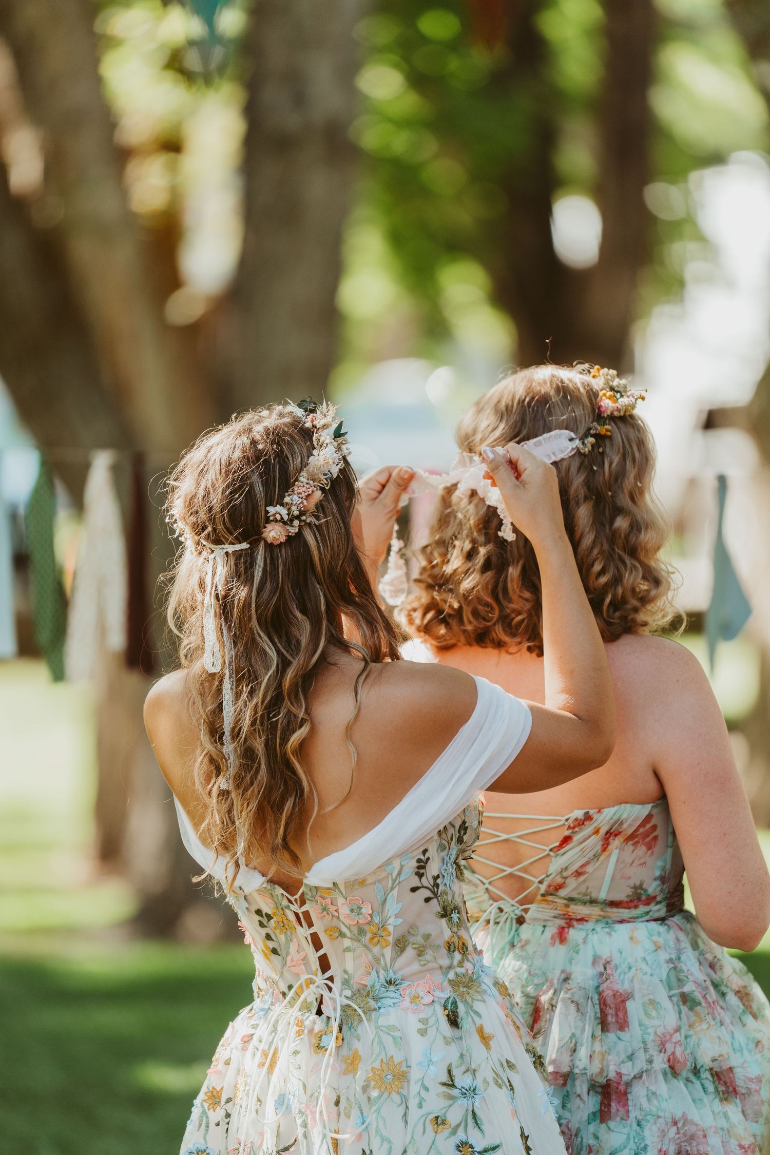 Two women in floral dresses and flower crowns stand outdoors among trees at a wedding celebration.