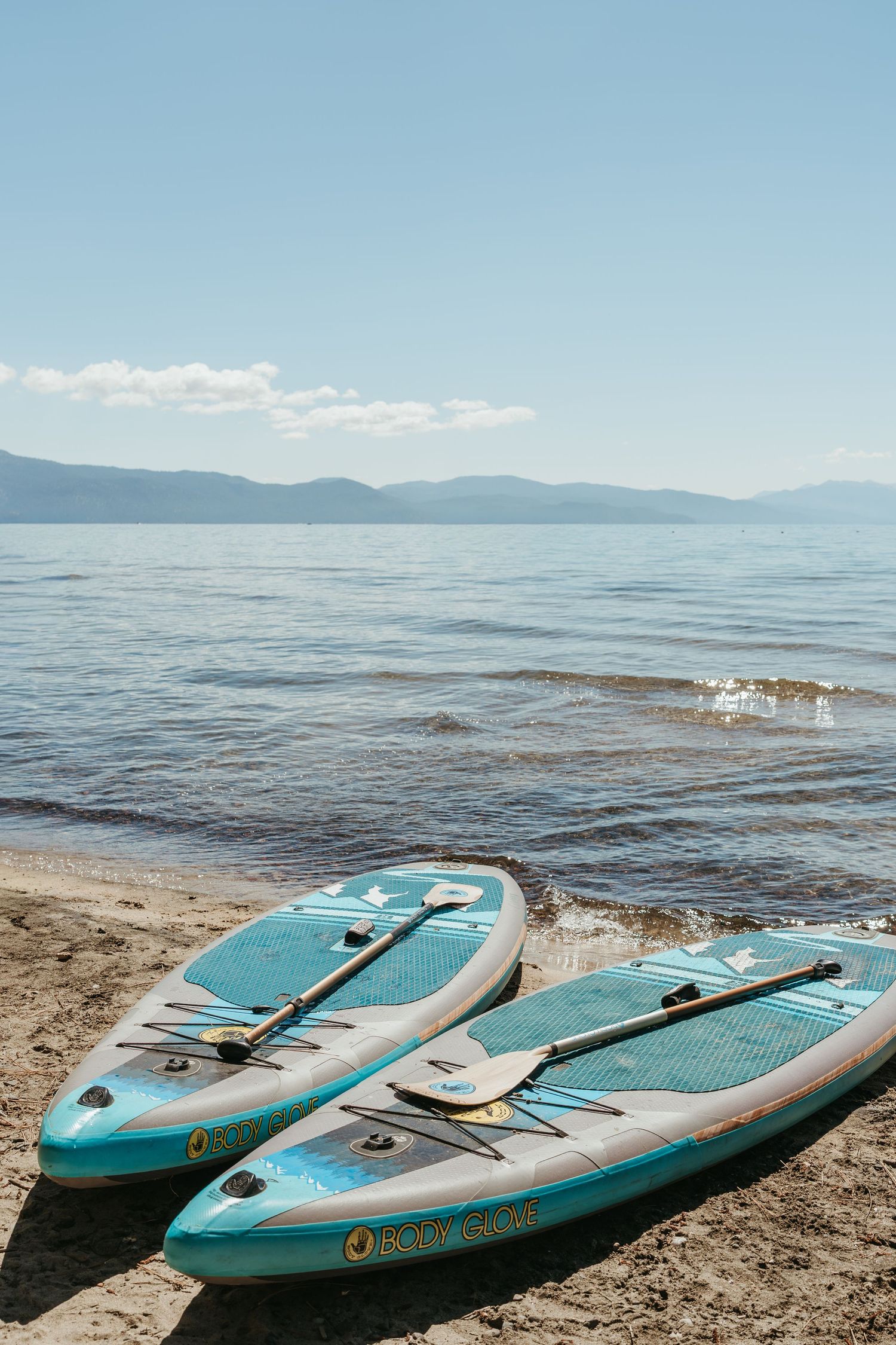 Two turquoise paddleboards rest on a sandy beach shore with mountains and calm lake waters in the background.