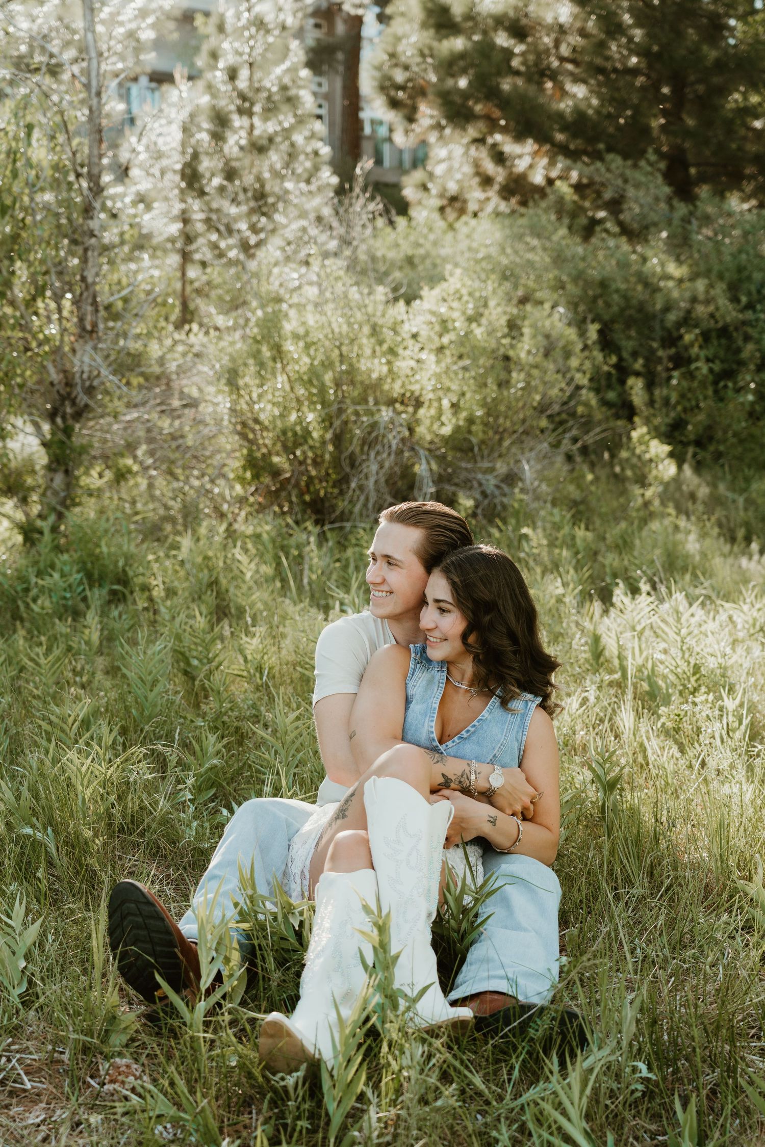 A couple sits together in a lush green meadow, smiling warmly, the woman wearing white cowboy boots and a denim outfit.