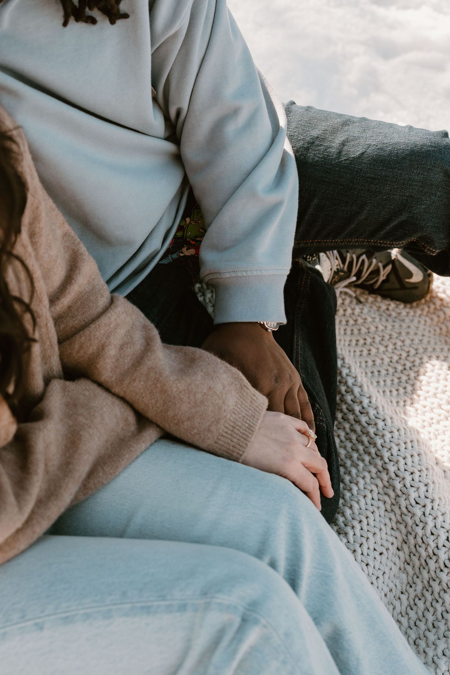 A couple sits closely together outdoors, holding hands, wearing cozy sweaters and jeans on a snowy winter day.