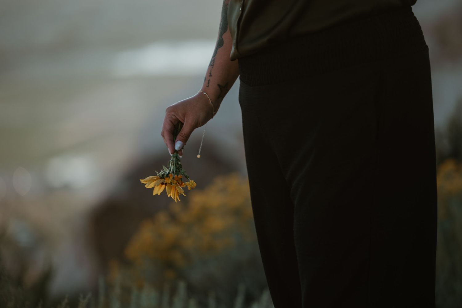 Person holding wilting yellow wildflowers at their side, wearing dark pants and olive shirt with tattoo visible.