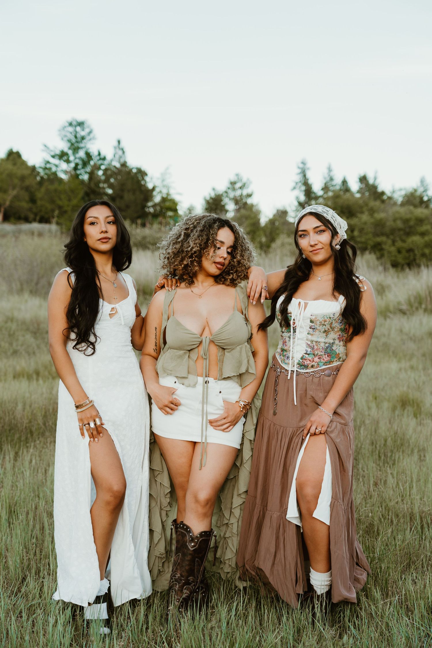 Three young women pose outdoors in a grassy field wearing boho summer outfits including corset tops and maxi skirts.