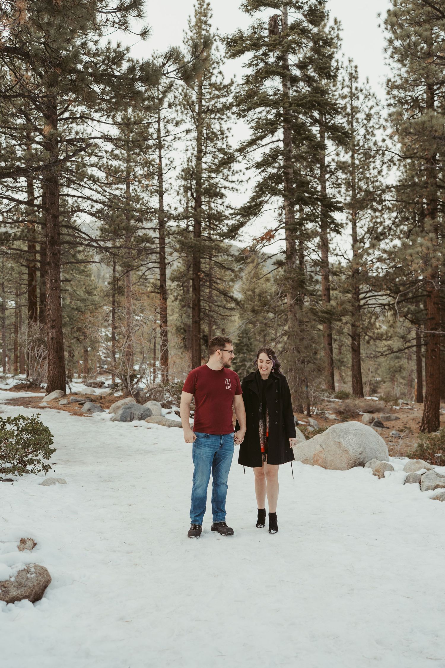 A couple stands together in a snowy winter forest surrounded by tall pine trees and scattered boulders.
