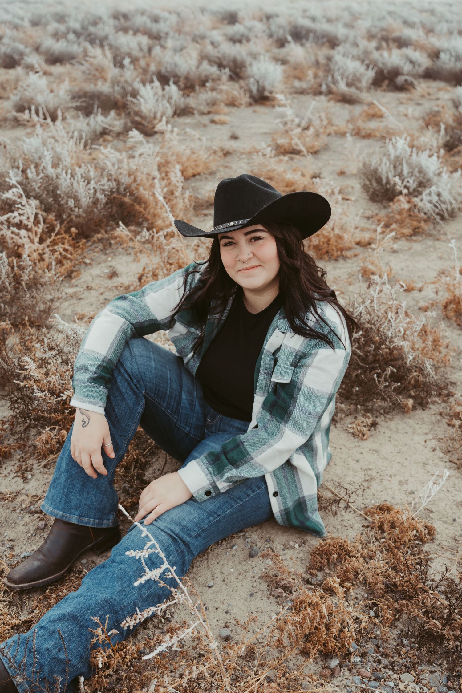 Western style portrait in desert landscape with cowboy hat, plaid jacket and distressed jeans against sage brush background.