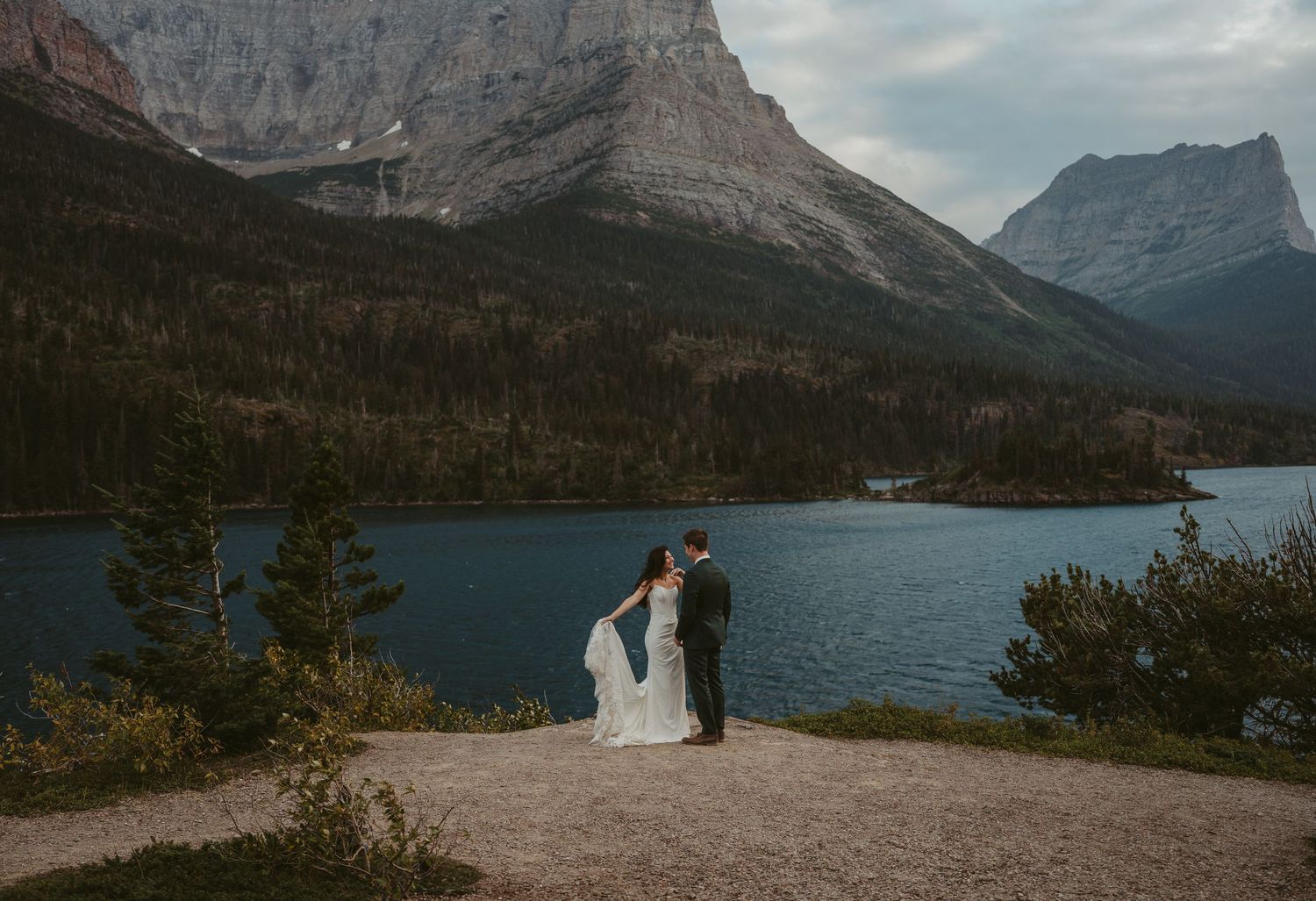 A couple shares a romantic moment on a scenic overlook above a mountain lake in Glacier National Park surrounded by towering peaks at dusk.
