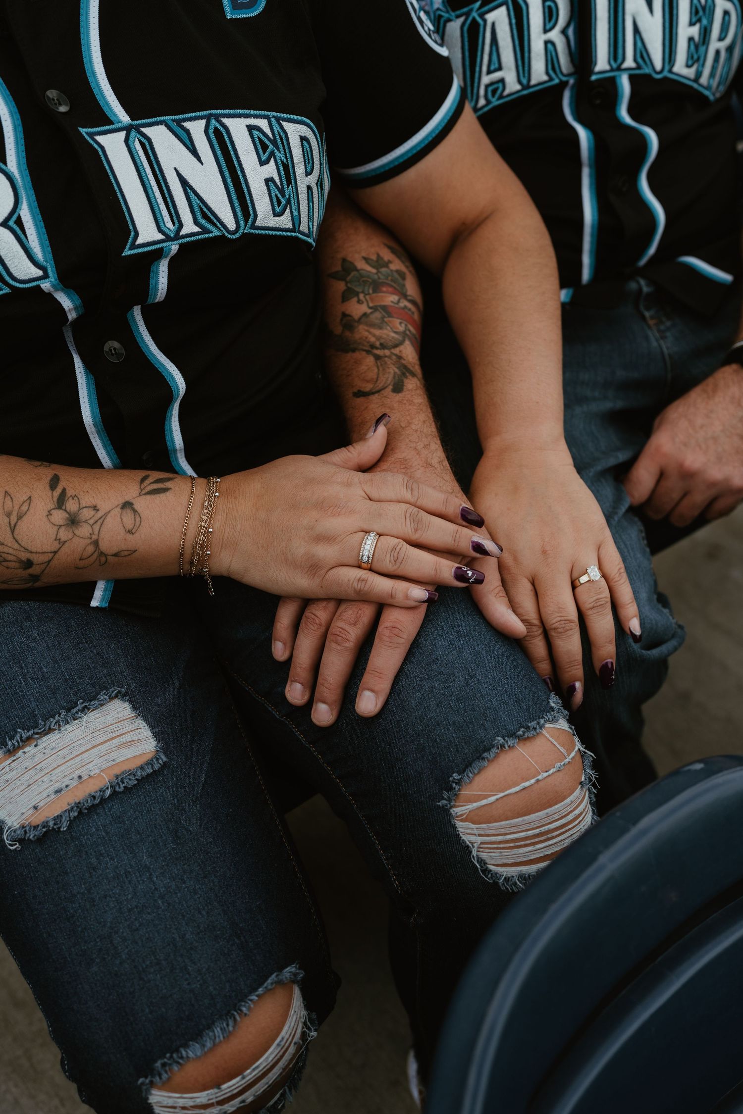 Couple wearing Seattle Mariners jerseys showing off wedding rings, with tattoos visible on their hands and arms.