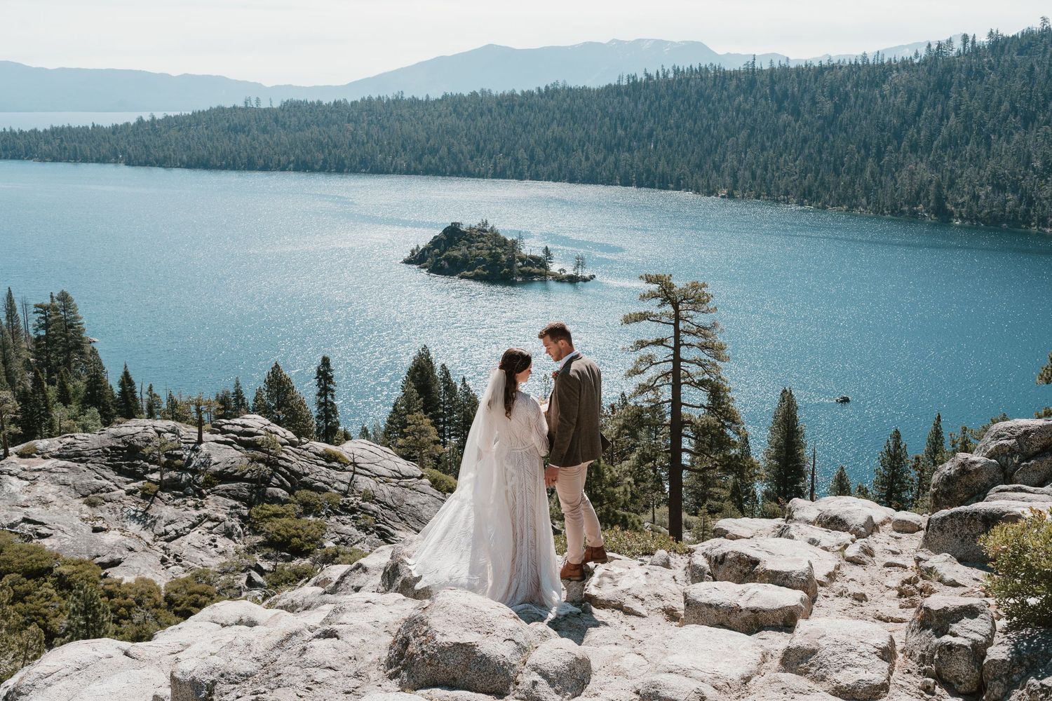 Bride and groom stand on rocky overlook above Emerald Bay at Lake Tahoe, surrounded by pine trees and blue water.