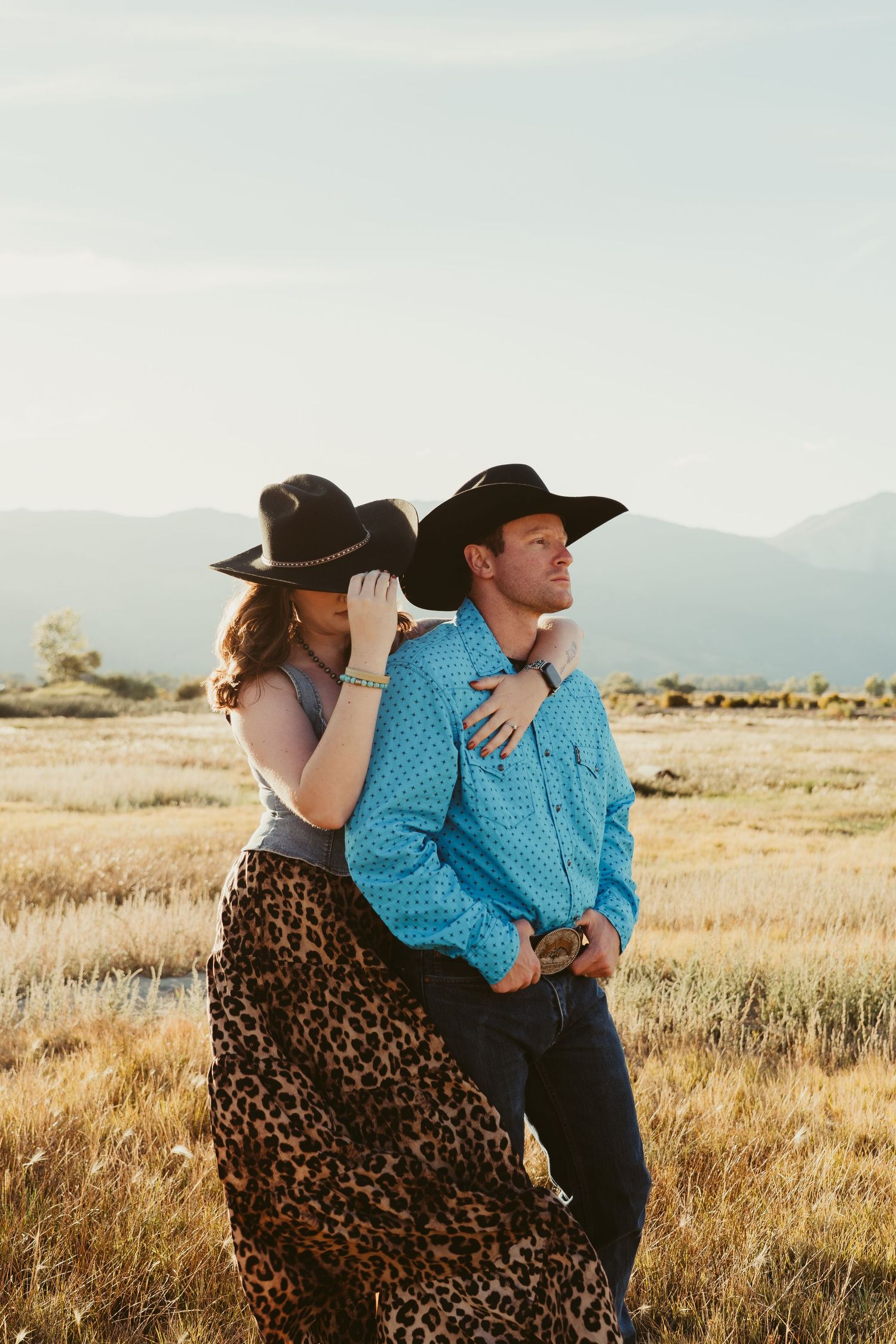 Couple in black cowboy hats embracing in a golden field with mountains, woman in leopard skirt, man in turquoise shirt.