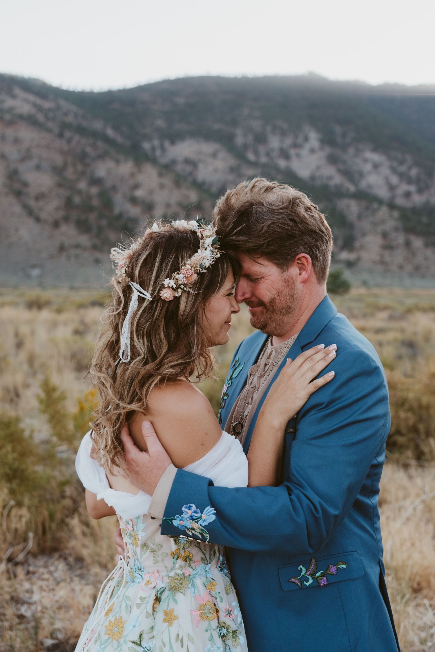 Couple in bohemian wedding attire embrace outdoors, bride in floral dress with flower crown, groom in blue suit, mountains behind.