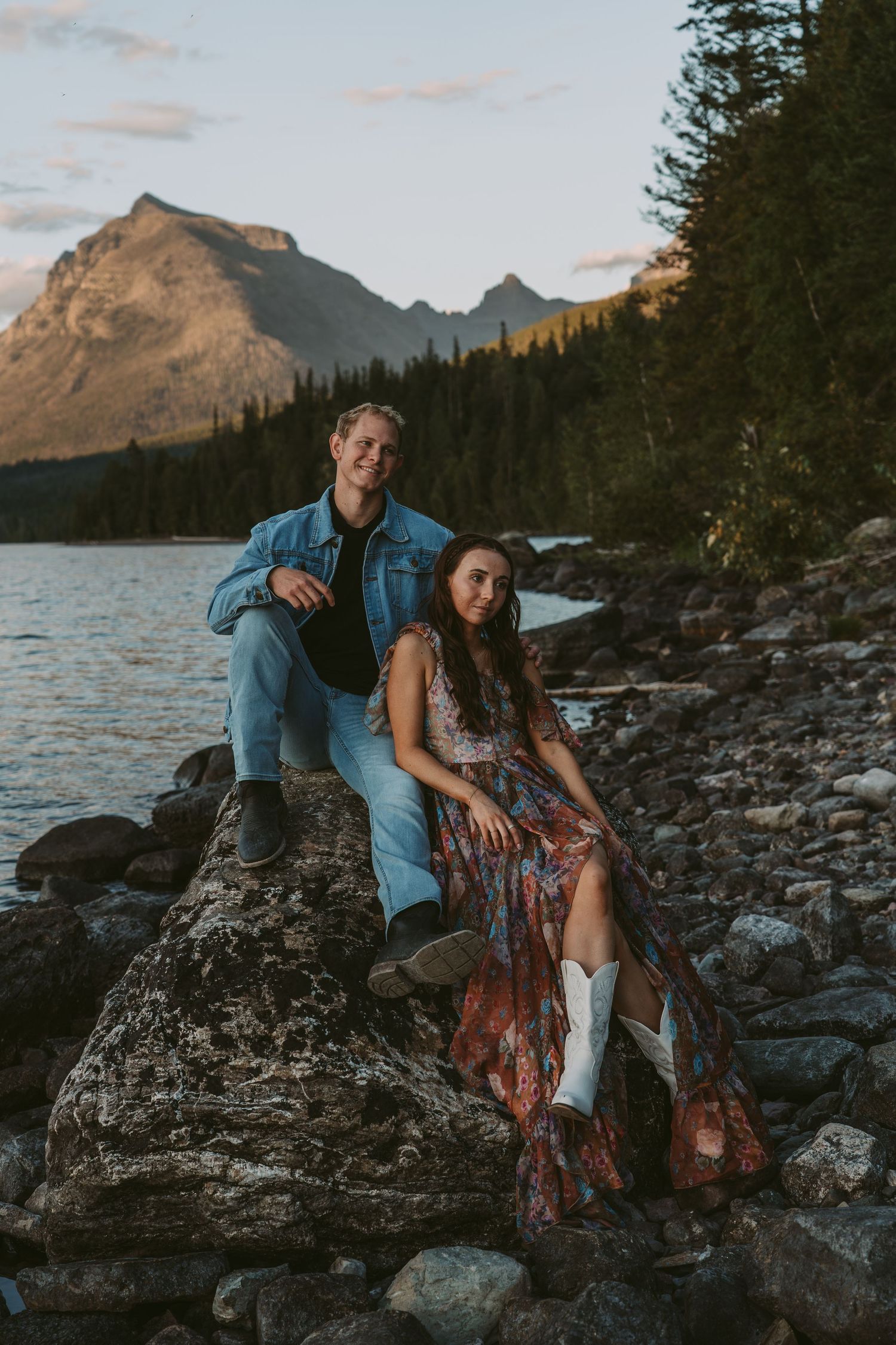 A couple poses together on rocky lakeside terrain with mountain peaks and evergreen forest in the background at sunset.