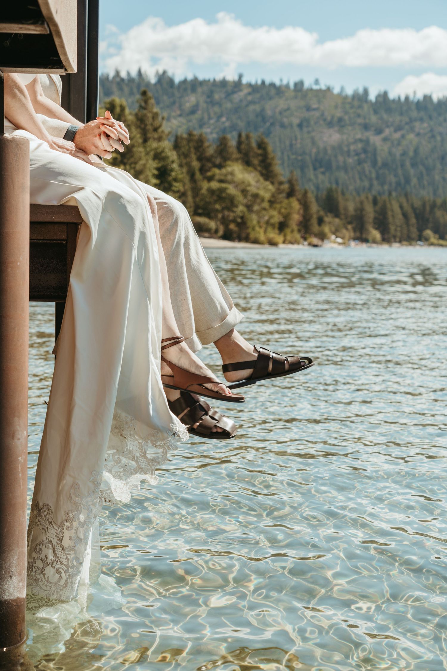 A person in white dress and brown shoes sits on a dock with feet dangling over sparkling lake water and forest backdrop.
